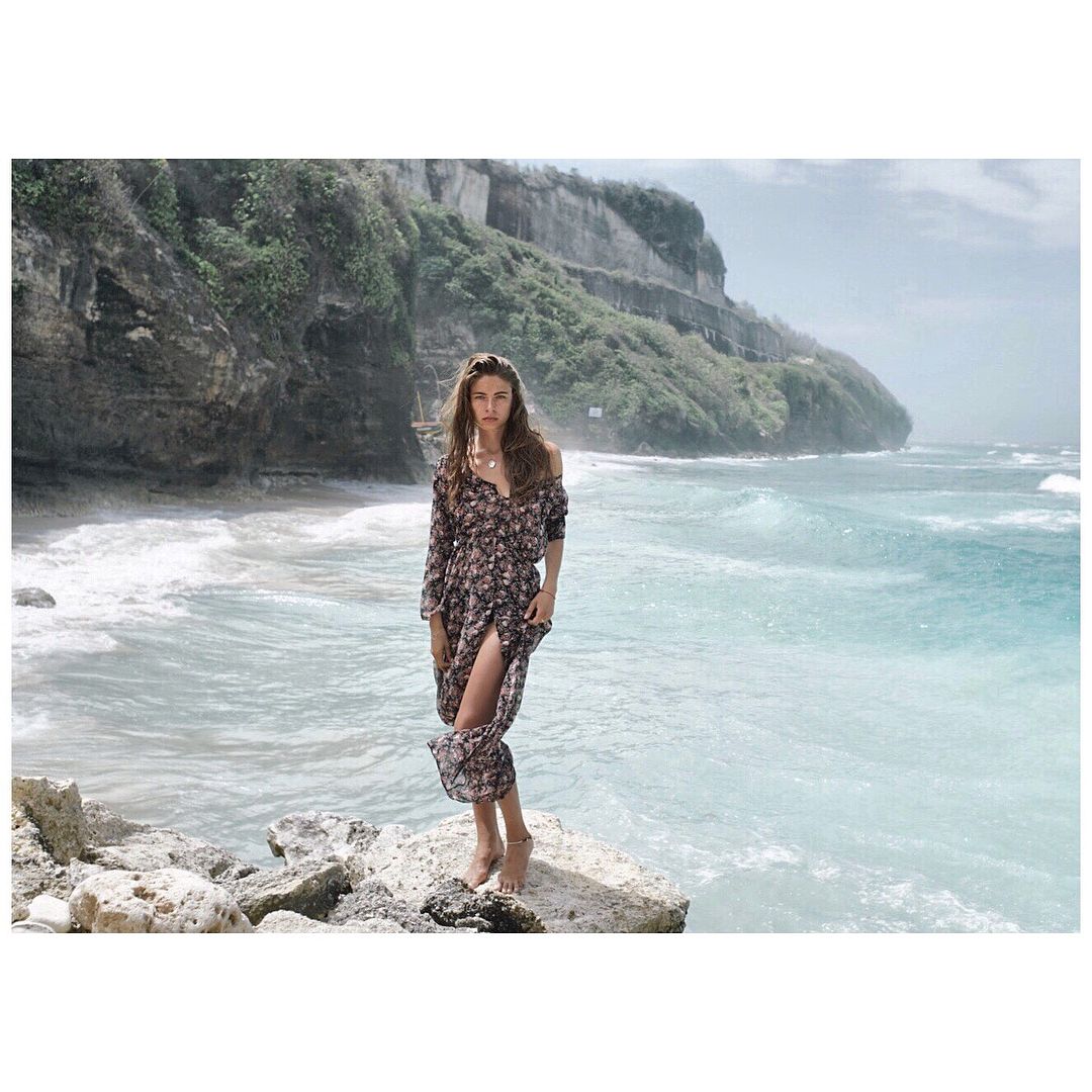 Woman in floral dress stands on a rock by the ocean with cliffs in the background.