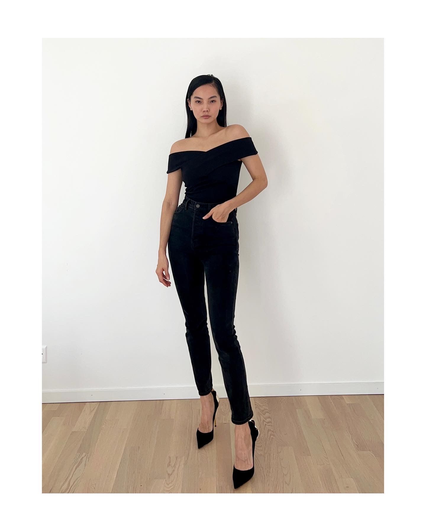 Woman in black off-the-shoulder top and jeans, hands in pockets, posing with black heels, against a white wall.