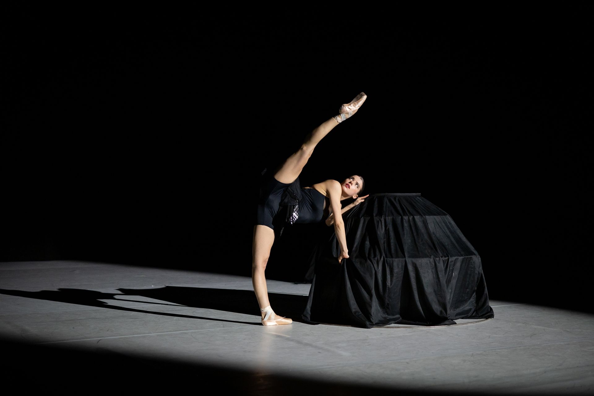 Ballet dancer balances, leg extended high, hand on covered structure, stage.