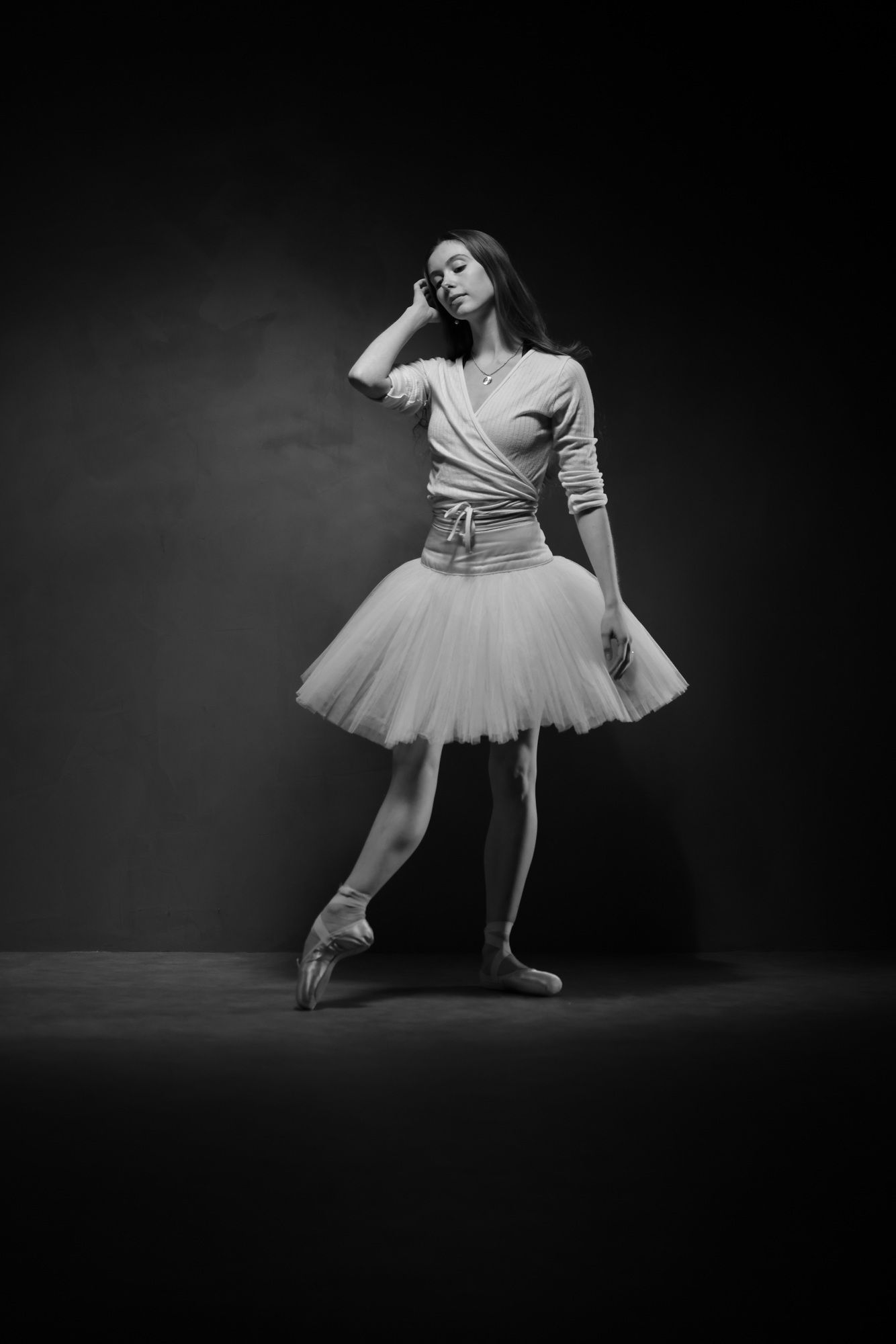 Ballerina in pointe shoes, tutu, and wrap sweater. Leaning on arm, dark background.
