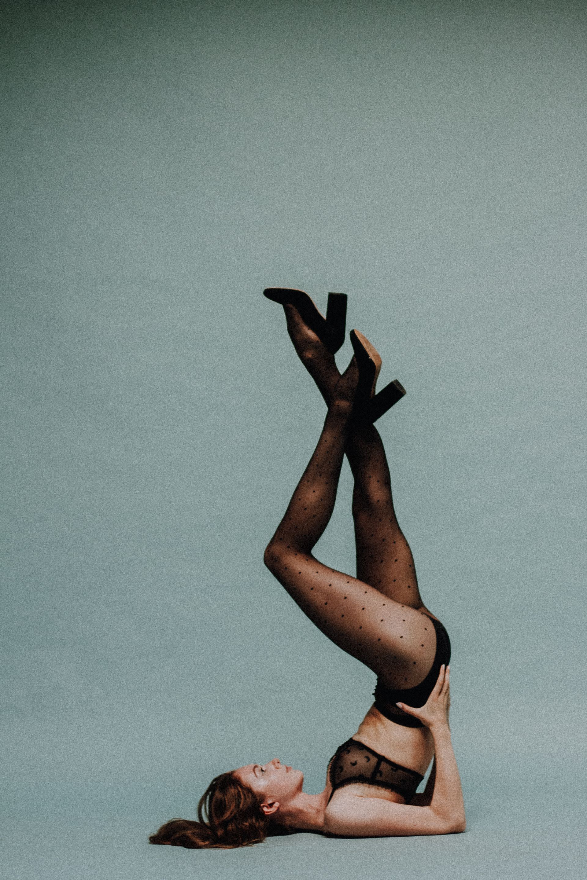 Woman in lingerie does a shoulder stand with legs crossed, wearing dotted tights and heels. Turquoise background.