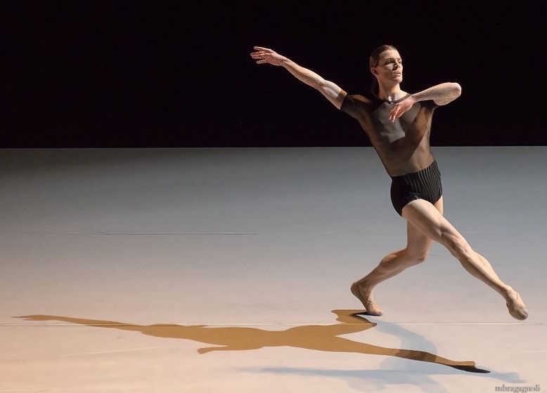 Ballet dancer in motion, on stage, dark top and shorts. Long shadow on the floor.