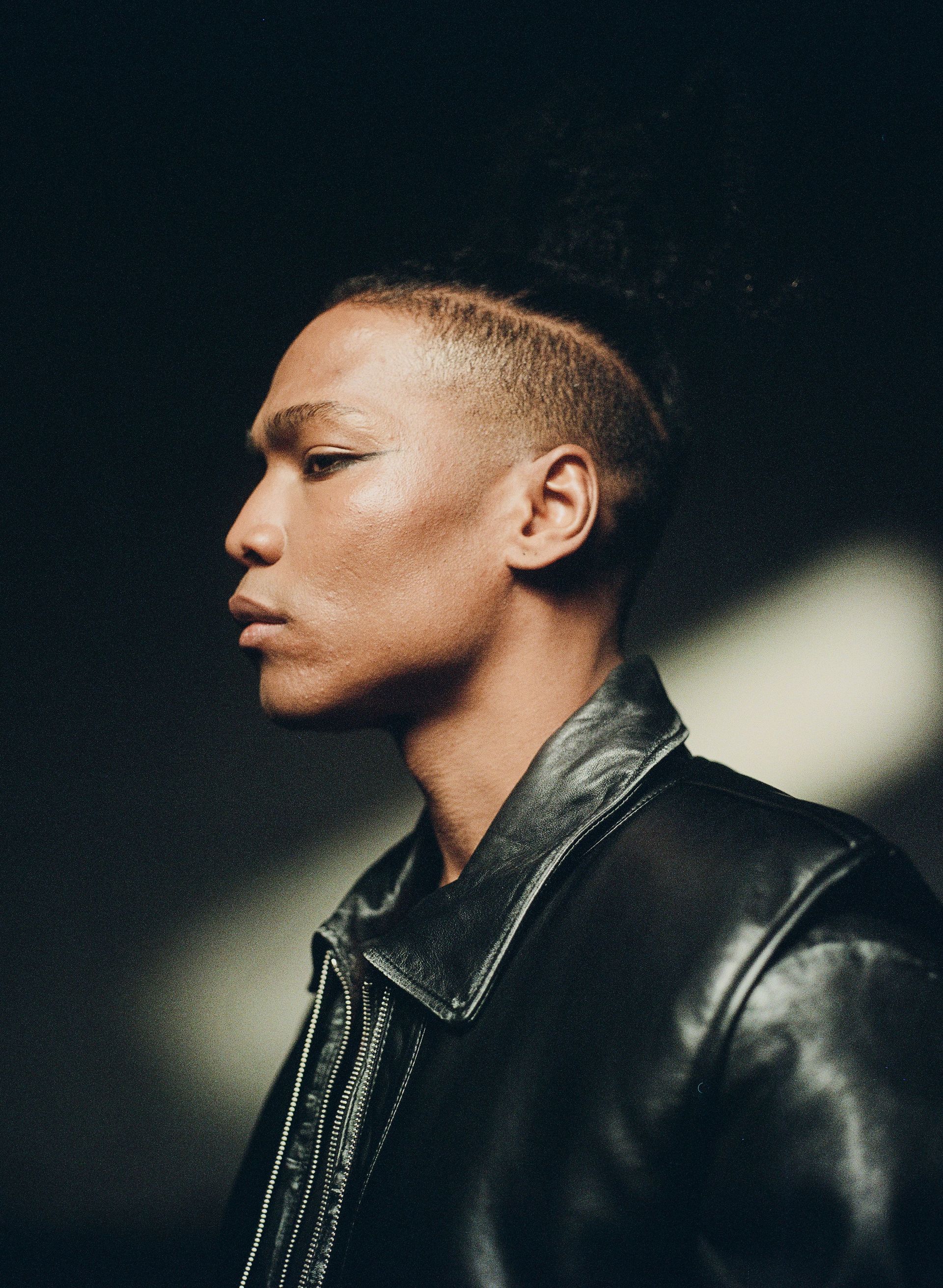 Person in a dark leather jacket, eyeliner, and shaved head, in profile. Dark background.