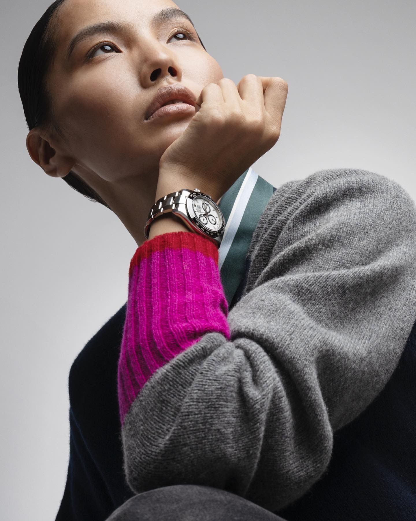 Person looking upward, wearing a watch. Hand on chin. Colorful sweater with pink cuff.