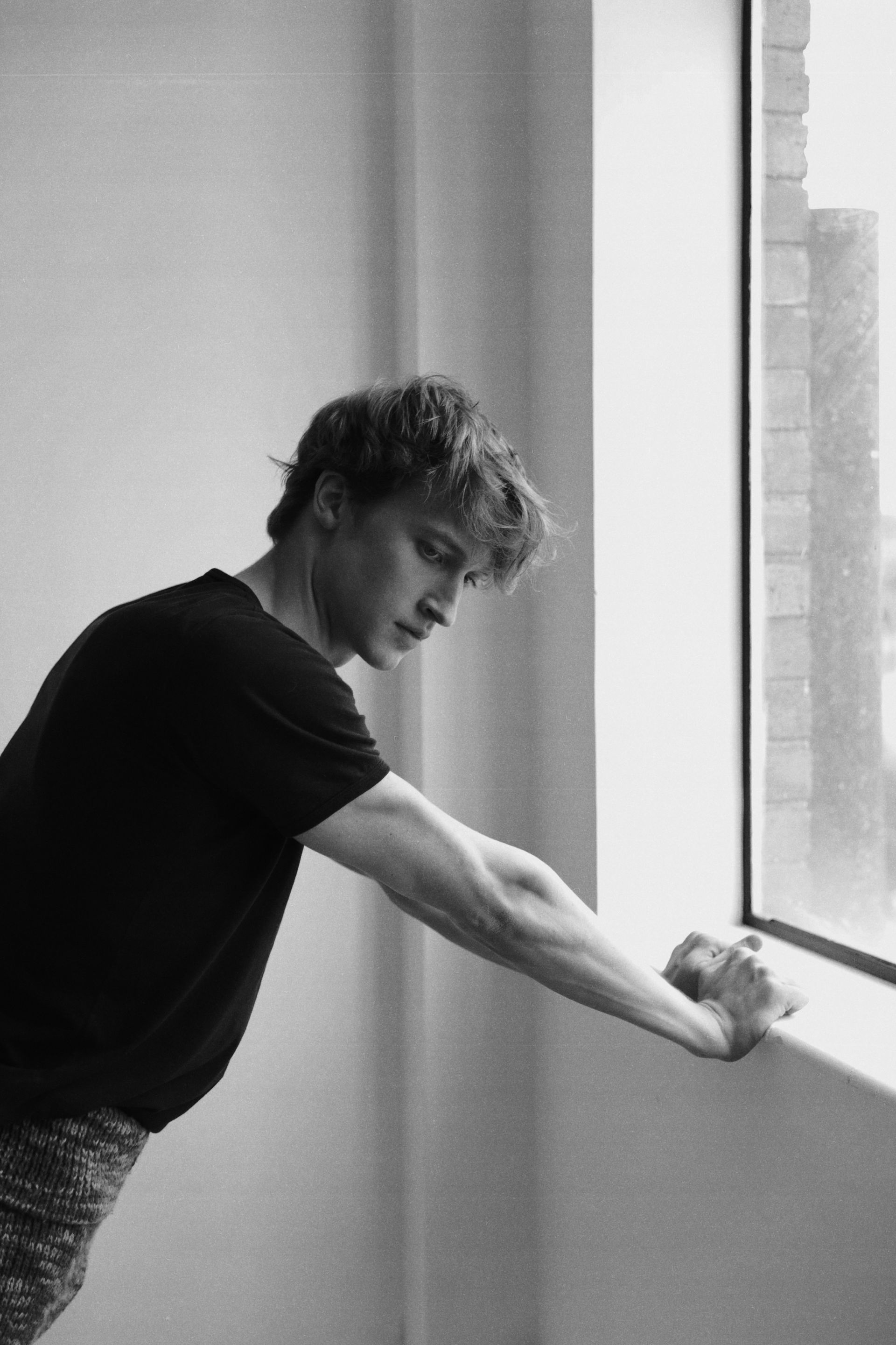 Man leaning against a window, arm extended. Black t-shirt, messy hair, indoors.