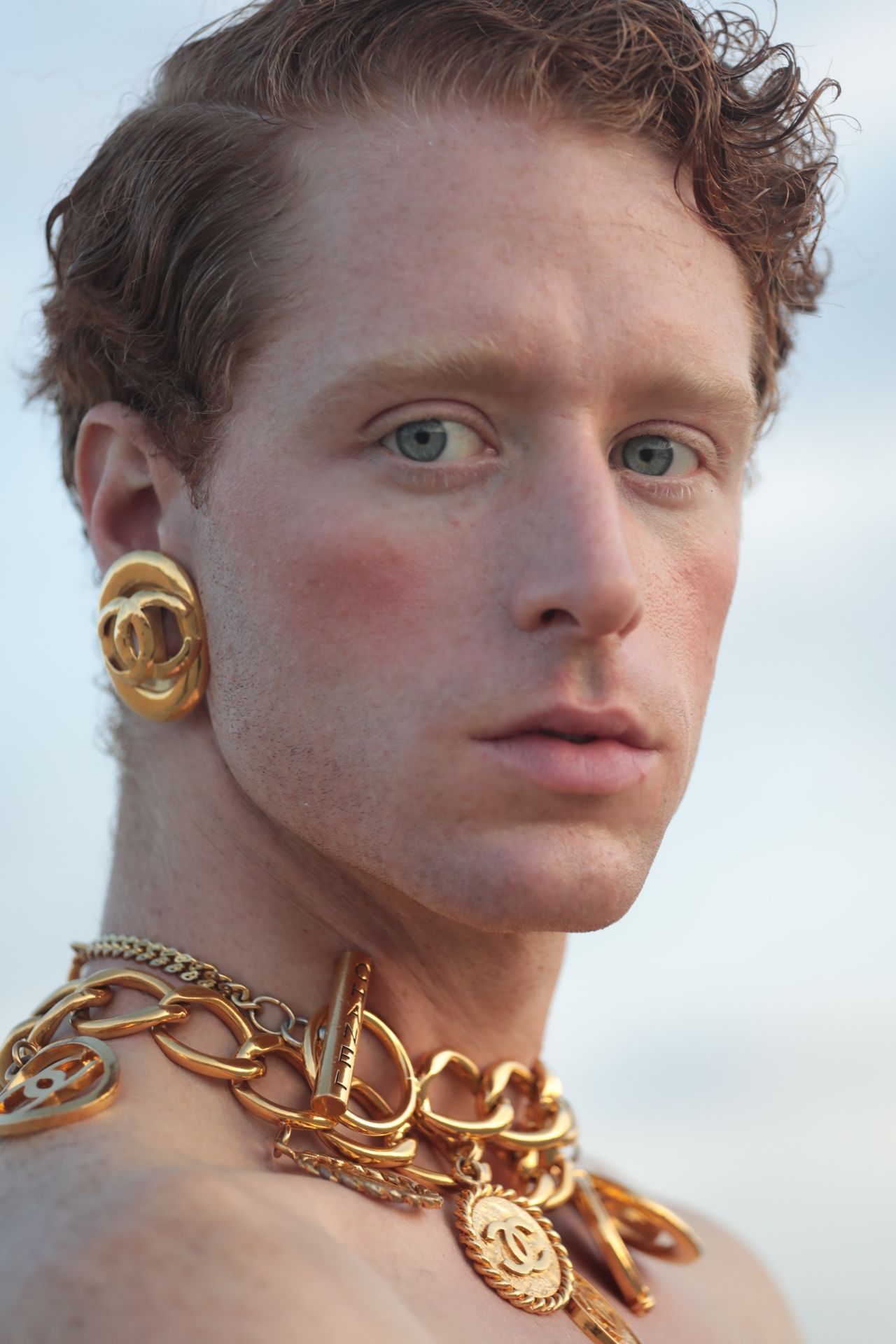Man with red hair, gold jewelry, and light blush, looking towards the camera against a blurred blue background.