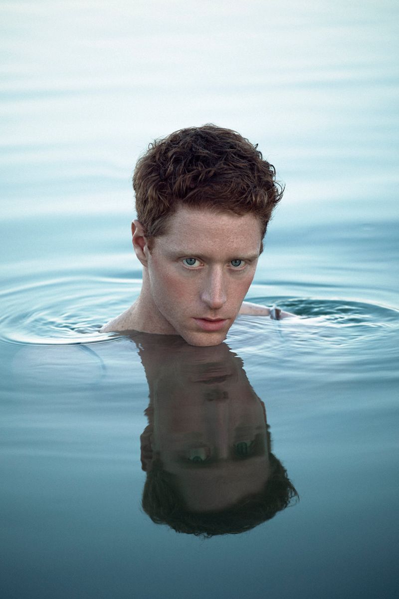 Man with red hair in water, reflected. Pale skin, gazing forward. Calm blue water.