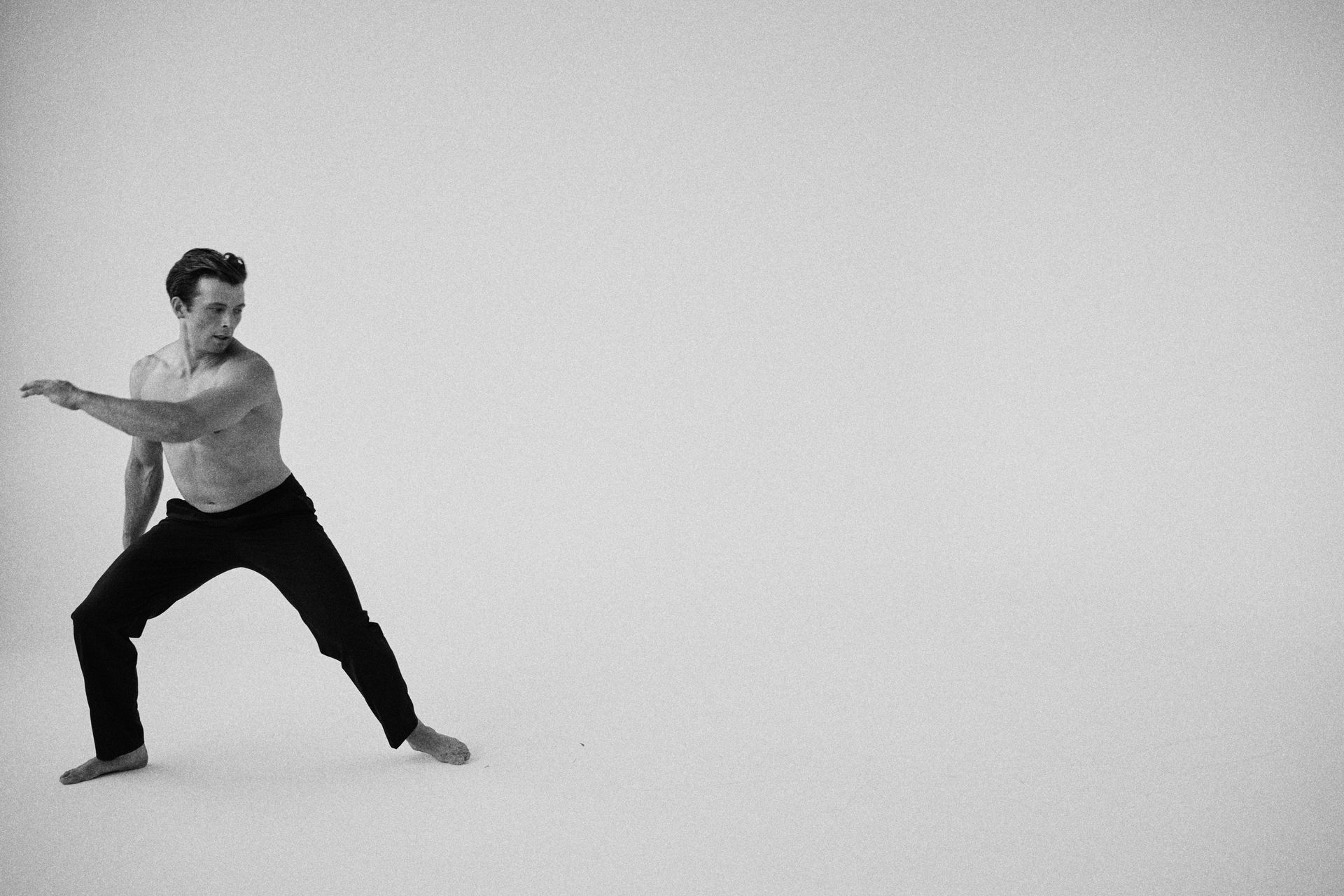 Man in athletic stance, shirtless, black pants, arm extended, white background.