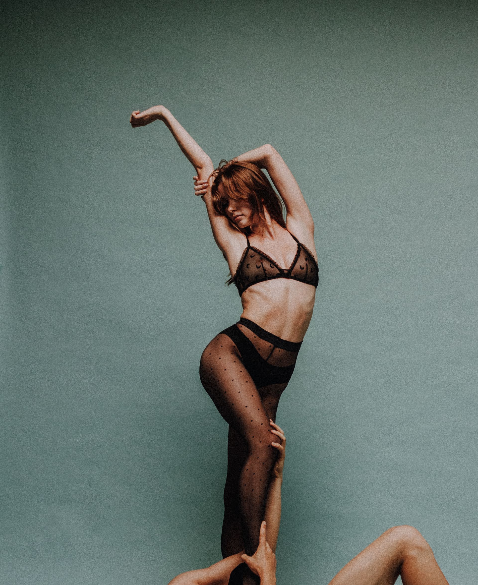 Woman in sheer lingerie being lifted with one arm raised against a teal background.