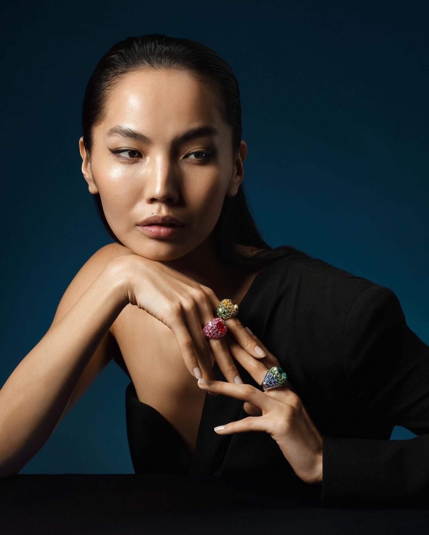 Woman with rings, resting chin on hands. Dark dress, jewel-toned rings against a blue backdrop.