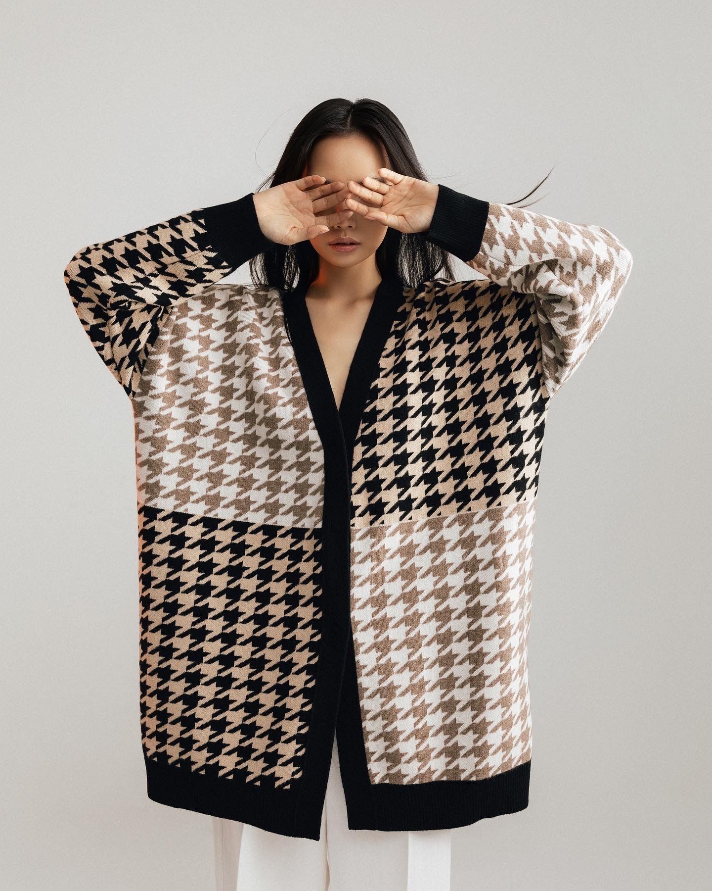 Woman in houndstooth cardigan covering eyes, standing in front of a white background.