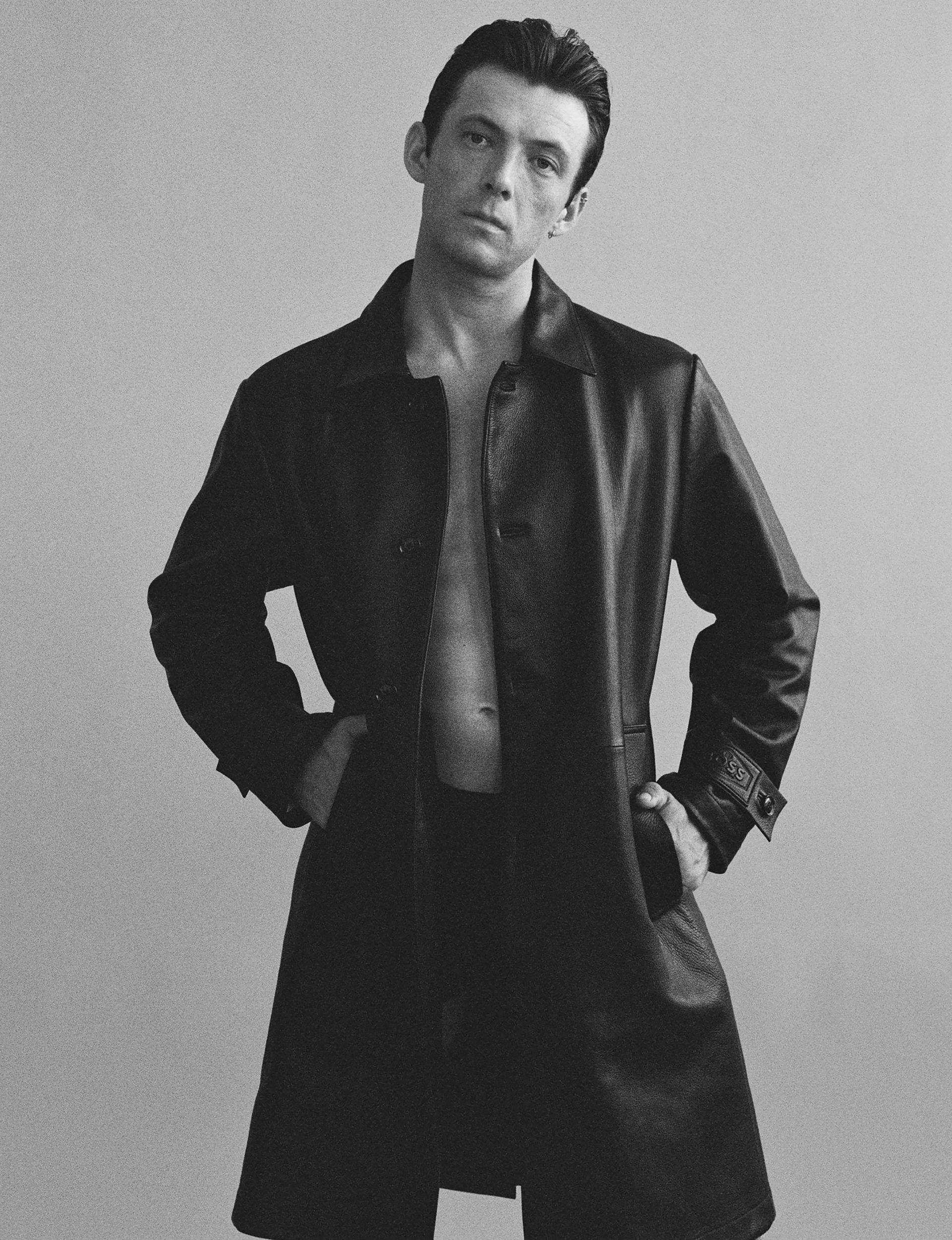 Man in long coat, hands in pockets, gazing at the viewer, shirt unbuttoned, monochromatic.