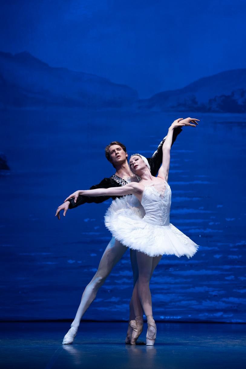 Ballet dancers performing on stage: woman in white tutu, man in black, both in graceful pose.