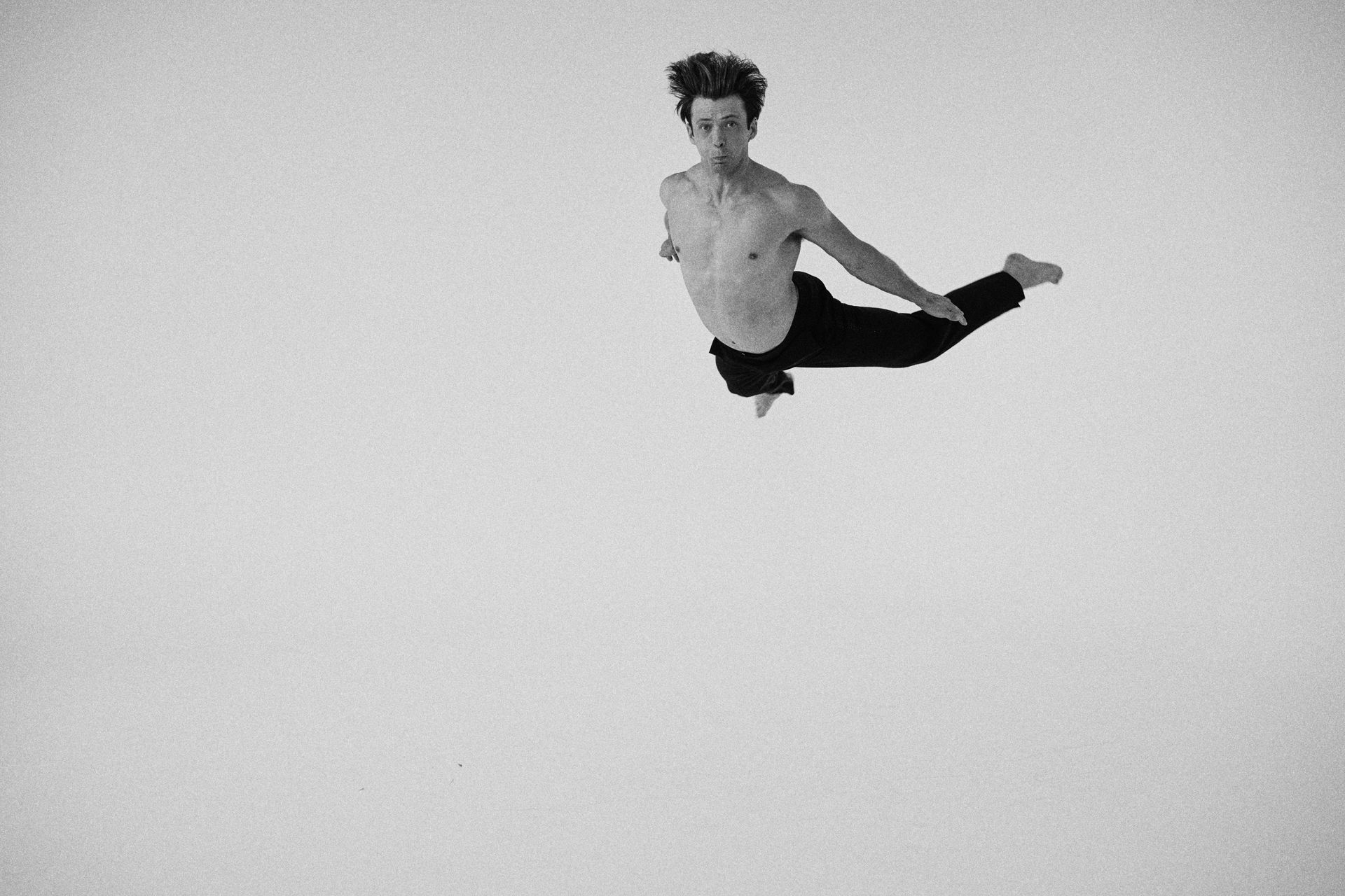 Man mid-air, leg extended. Wearing black pants, shirtless. Gray background.