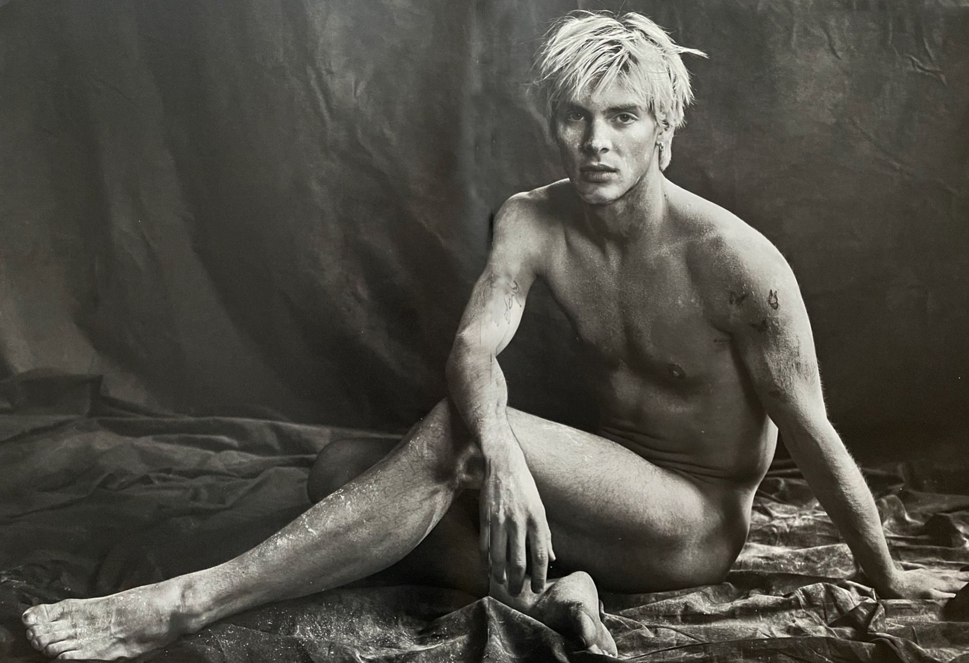 Nude person sitting, covered in substance, looking at camera. Blonde hair, dark background.