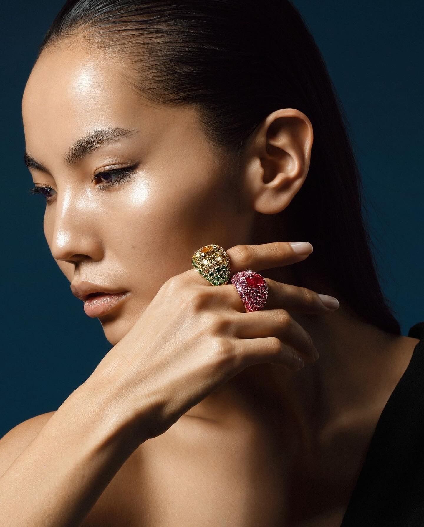 Woman with sleek hair, wearing jeweled rings, hand near face, blue background.