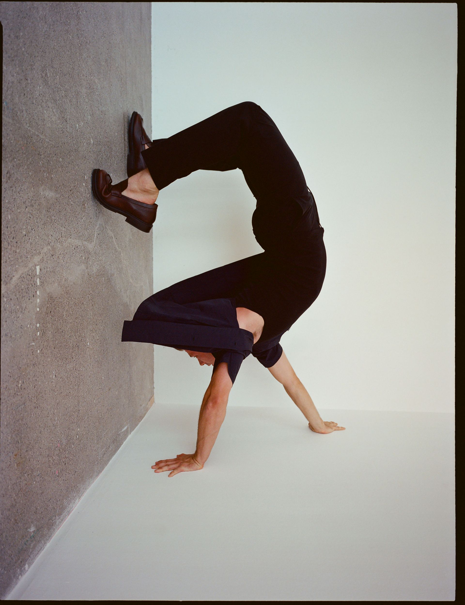 Person in black clothing doing a handstand in the corner of a room, feet against the wall.