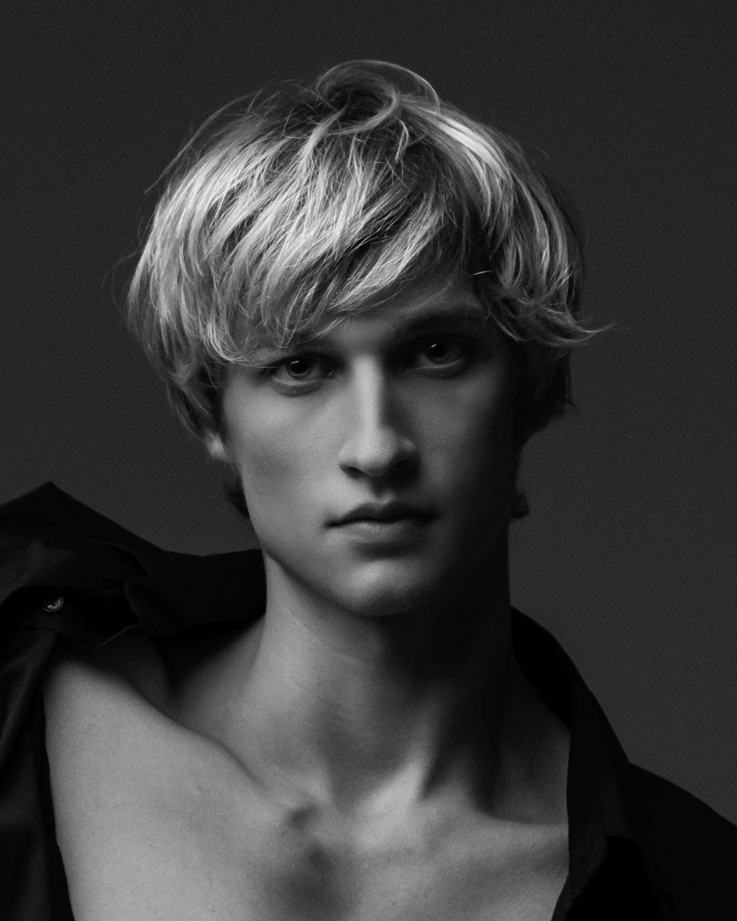 Blond-haired person looking directly at the viewer with a neutral expression, dark shirt off the shoulder.