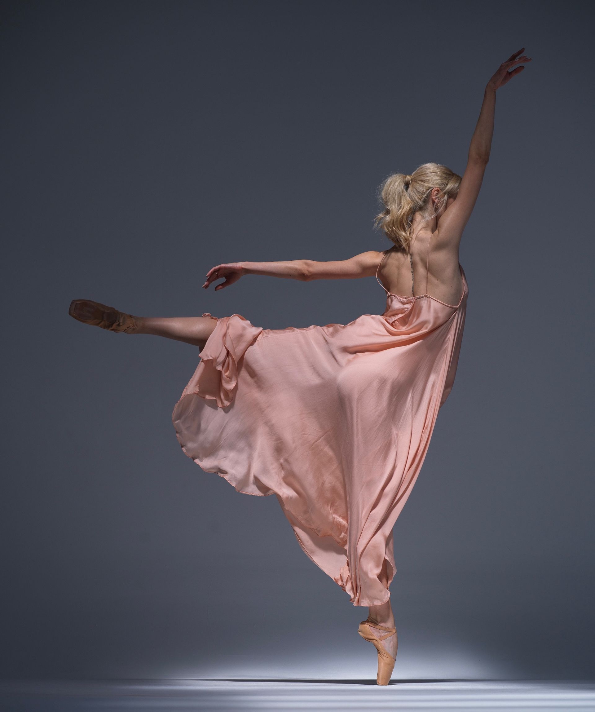 Ballerina in peach dress en pointe, leg extended, arms gracefully posed on a gray background.