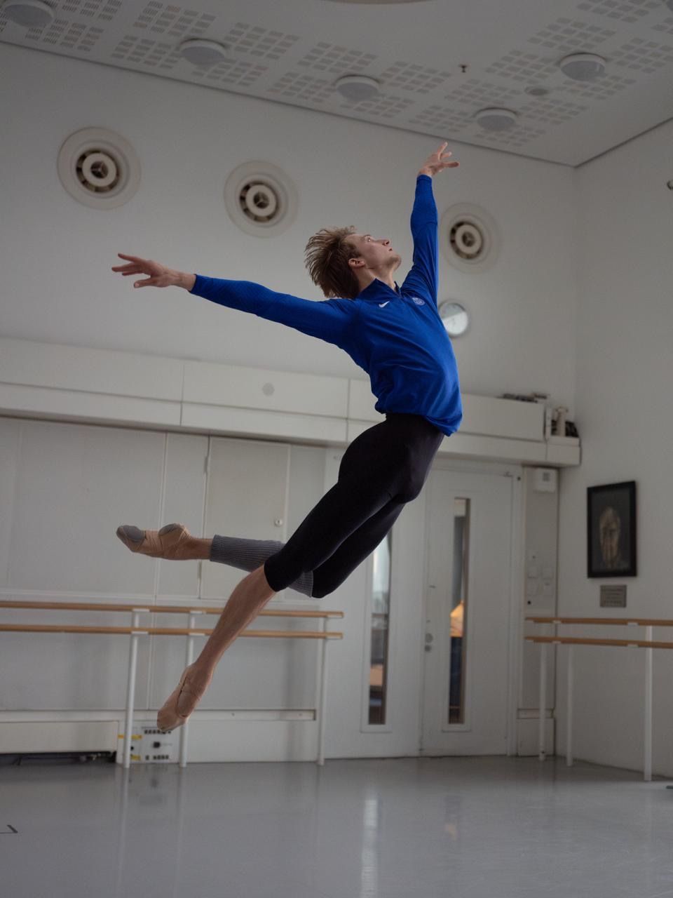 Male ballet dancer in mid-air leap, arms outstretched, wearing blue jacket and black tights, in a dance studio.