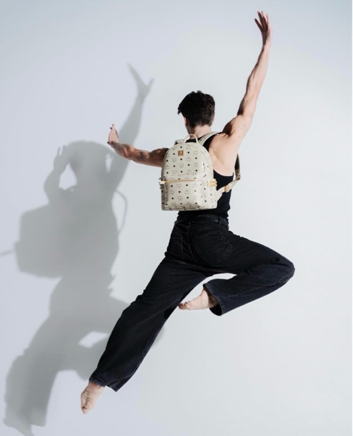 Person leaping in the air, wearing a black top, jeans, and backpack with a white background.