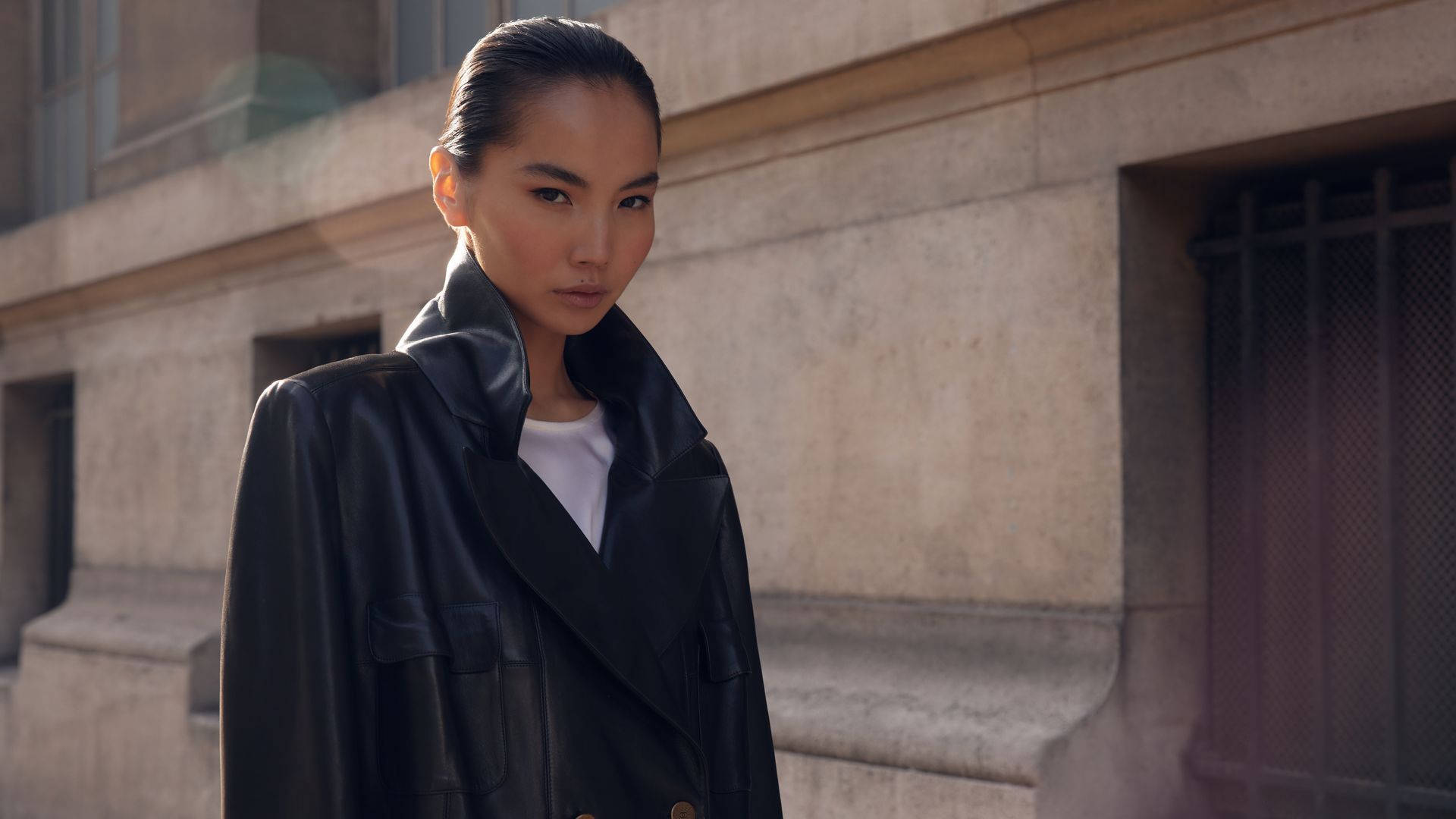 Woman in black coat, standing outdoors in front of a building, looking towards the camera.