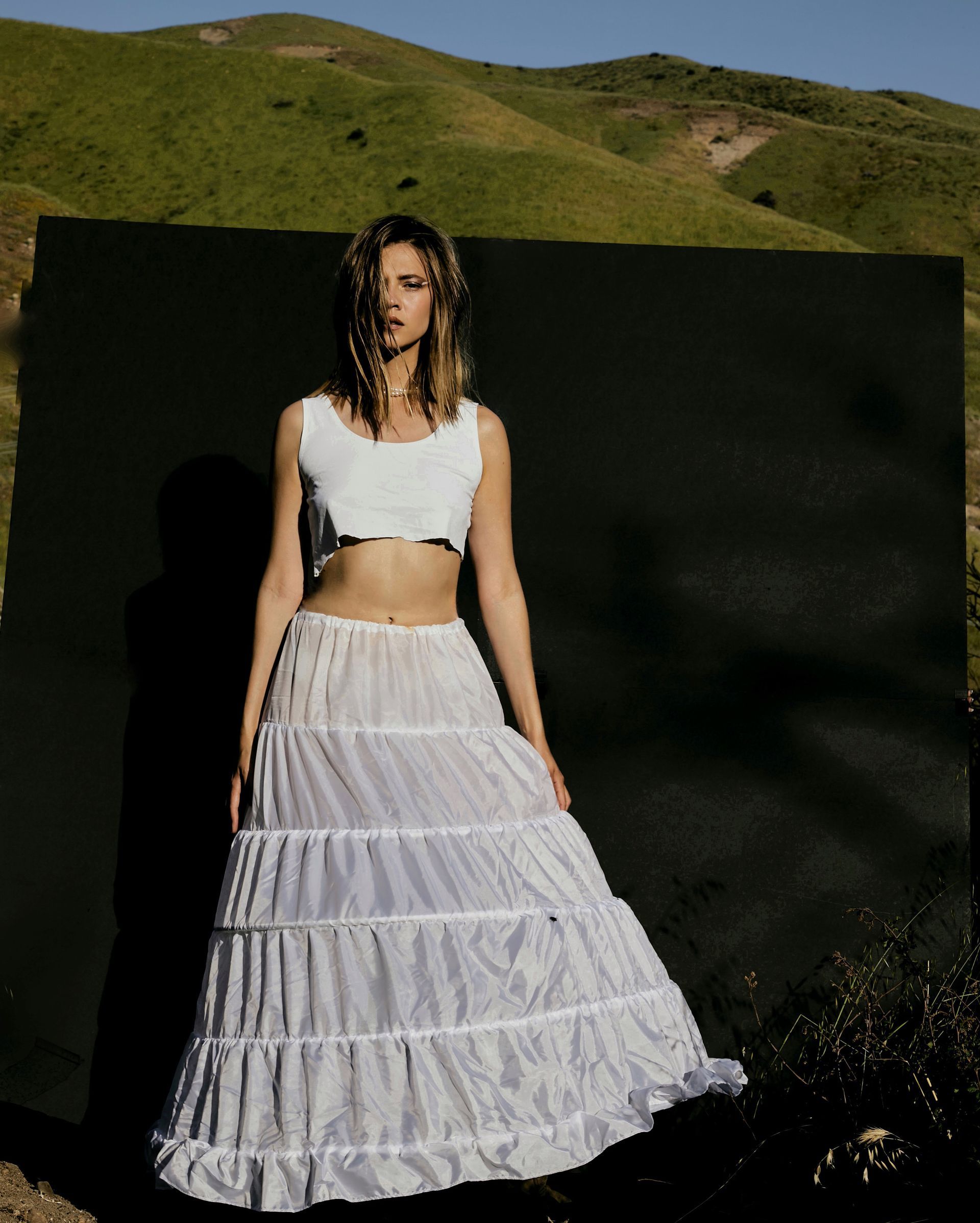 Woman in crop top and tiered skirt poses in front of a black backdrop with mountains in the background.