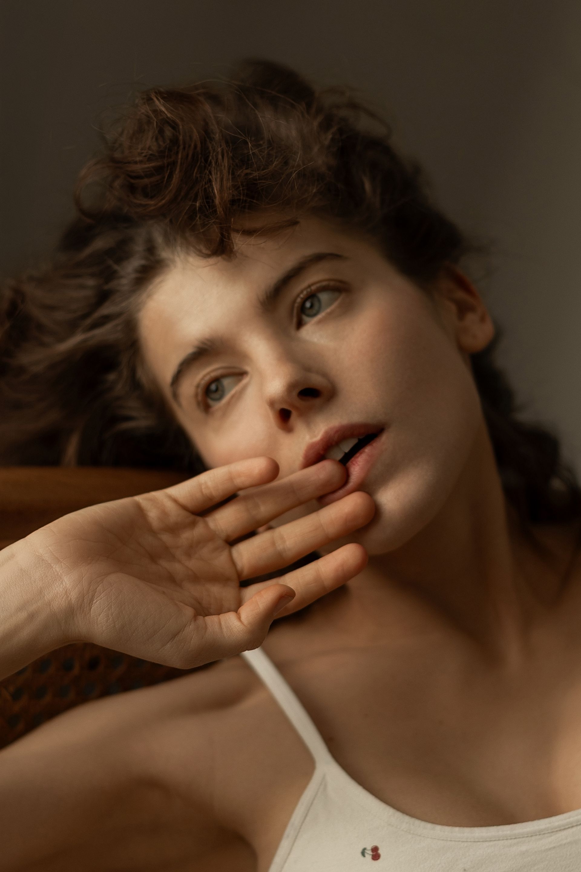 Woman biting a finger, looking up pensively.  Wearing a white tank top, sitting indoors.