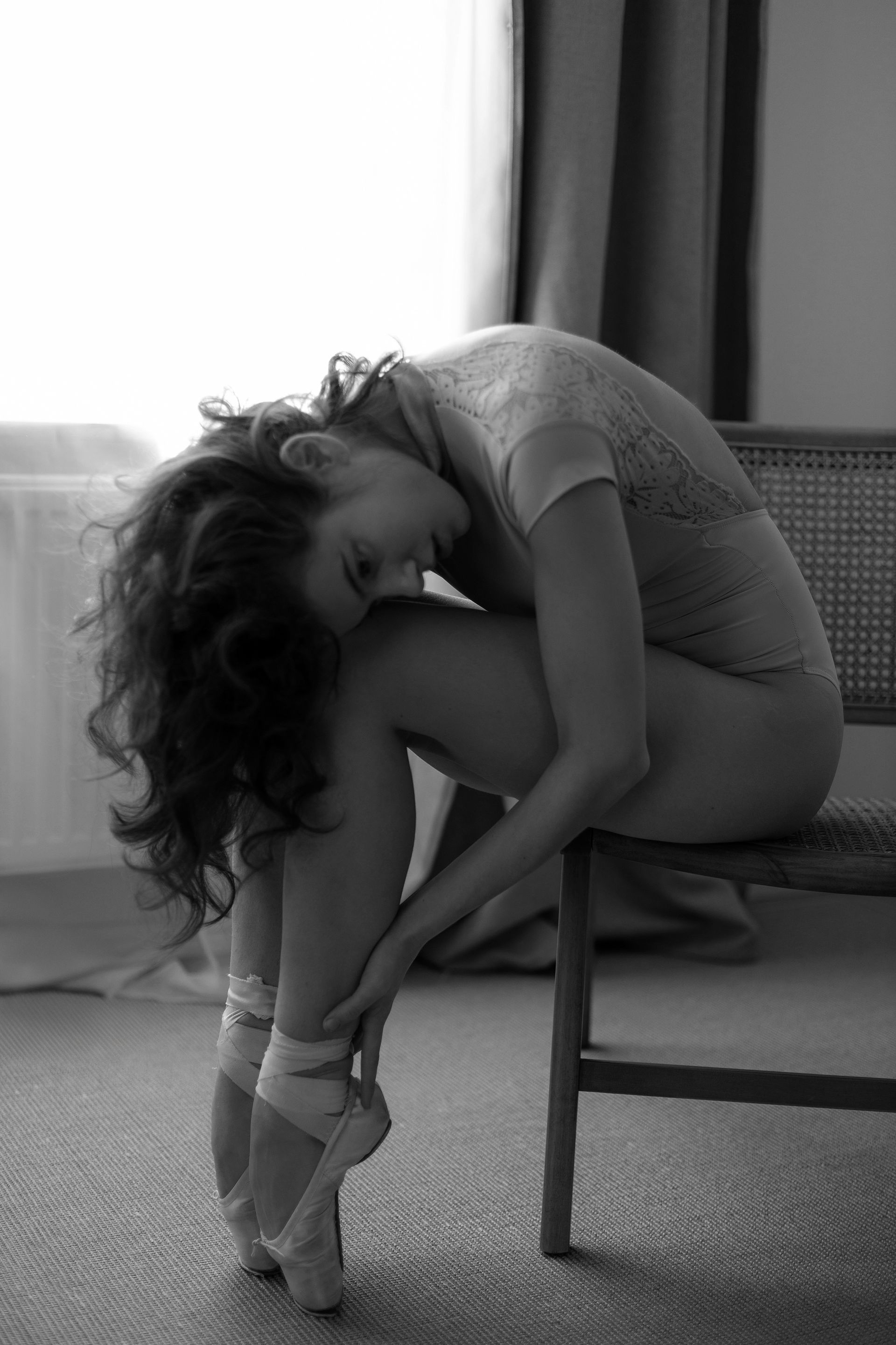 Ballerina in pointe shoes sits on a chair, head resting on her knees, indoors in black and white.