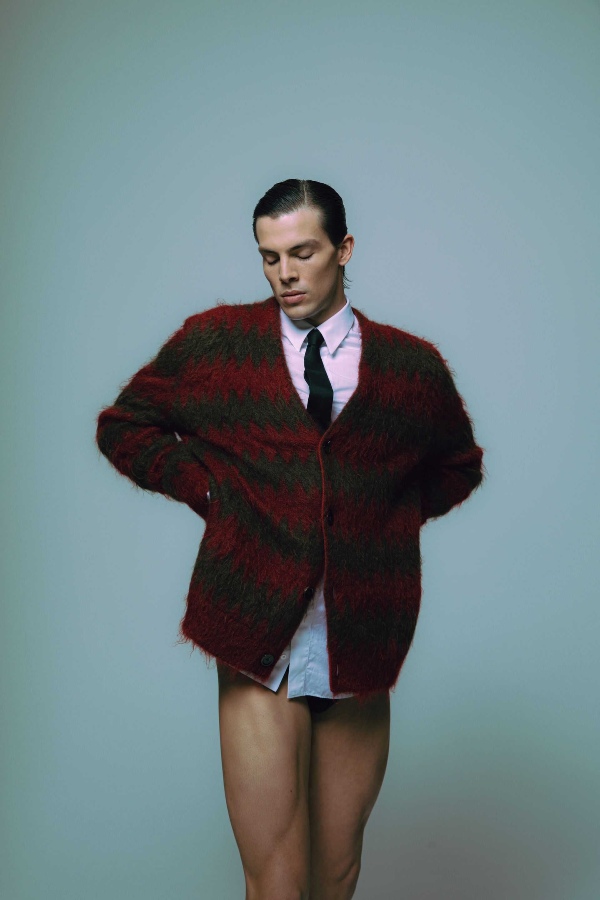 Man in oversized striped cardigan, tie, and underwear, with hands at waist, set against a plain backdrop.