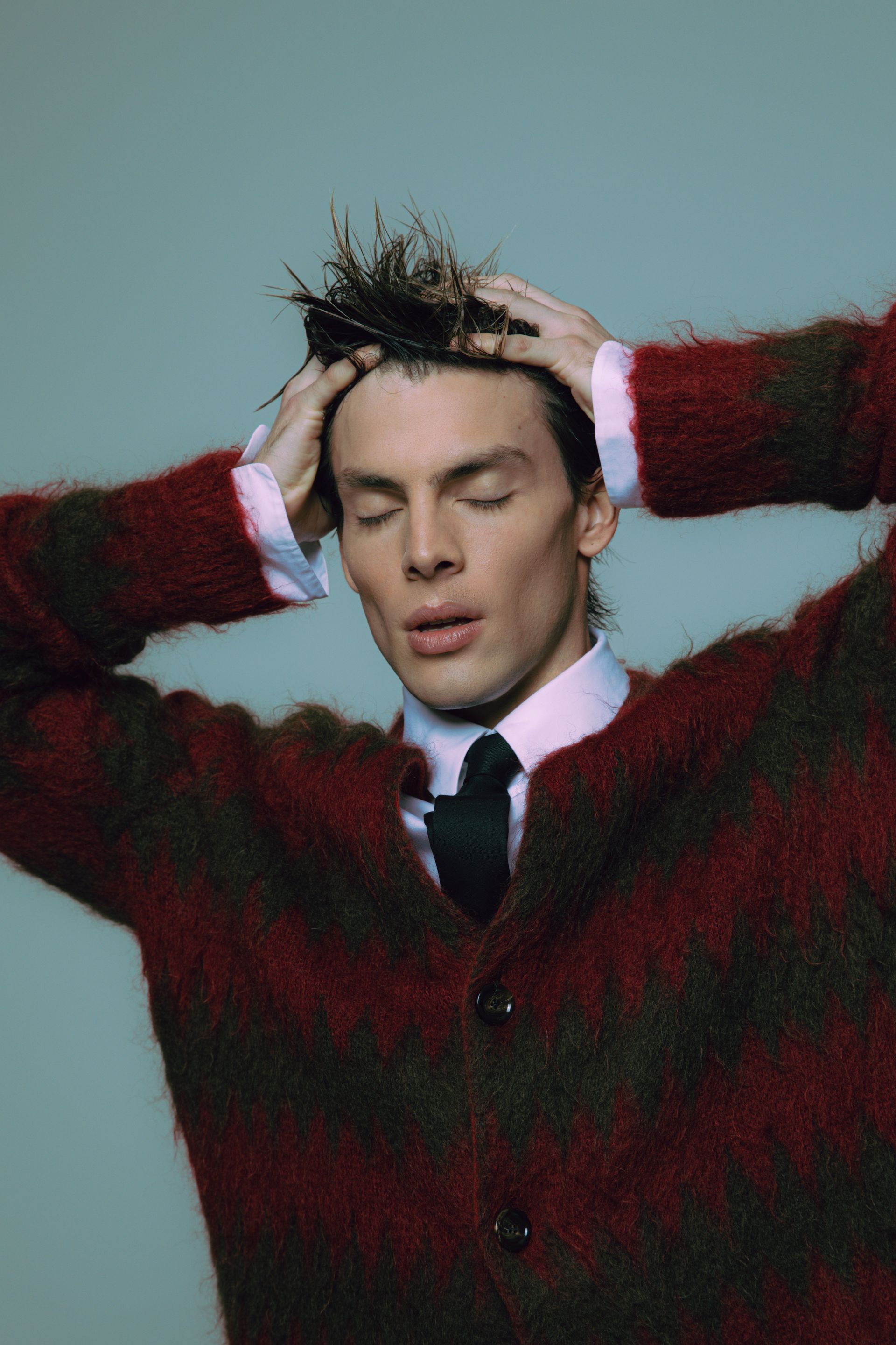 Man in red and brown striped sweater, eyes closed, hands on head.