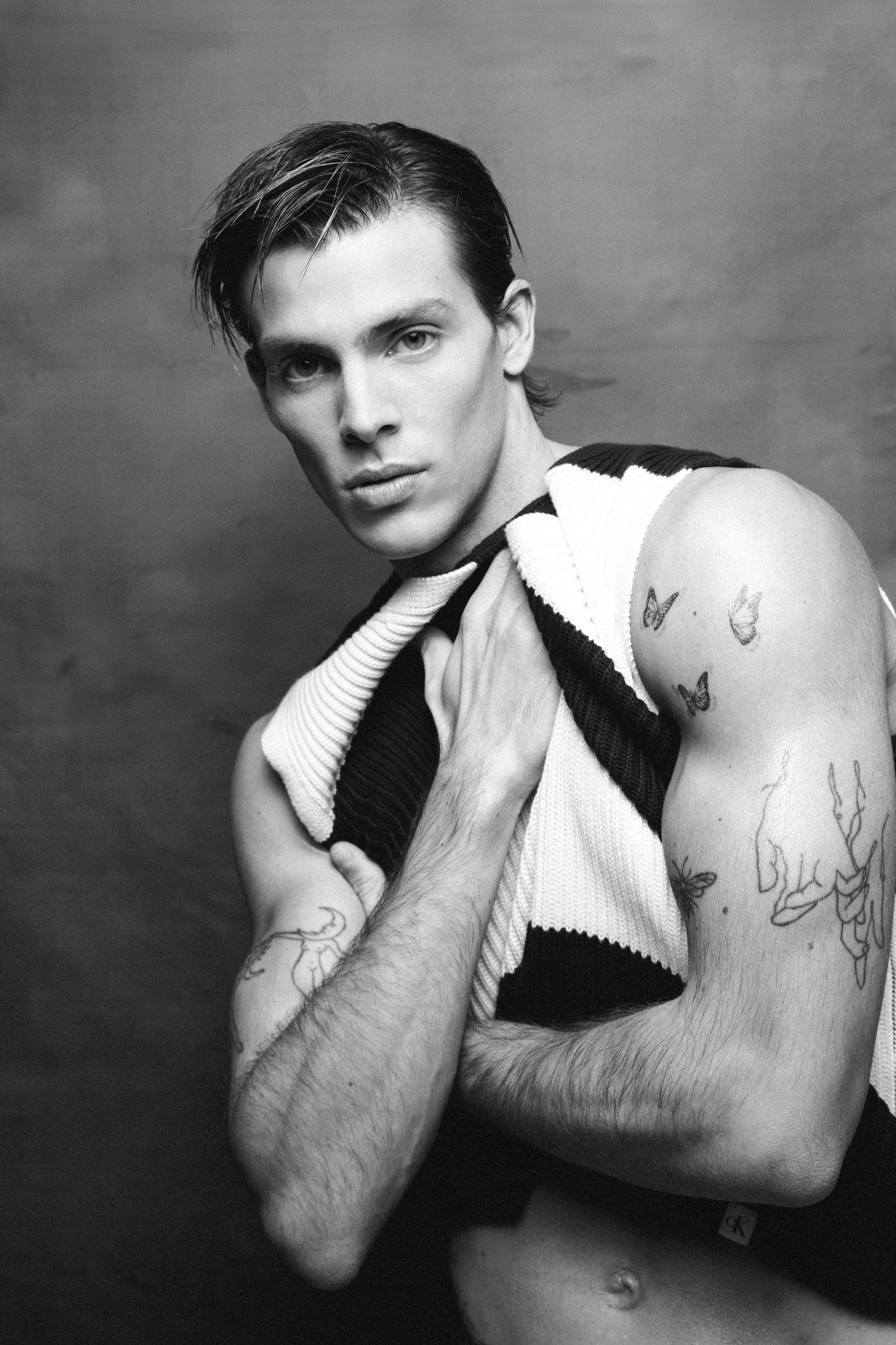 Man with tattoos, holding a black and white striped garment, looking at the camera with a serious expression.