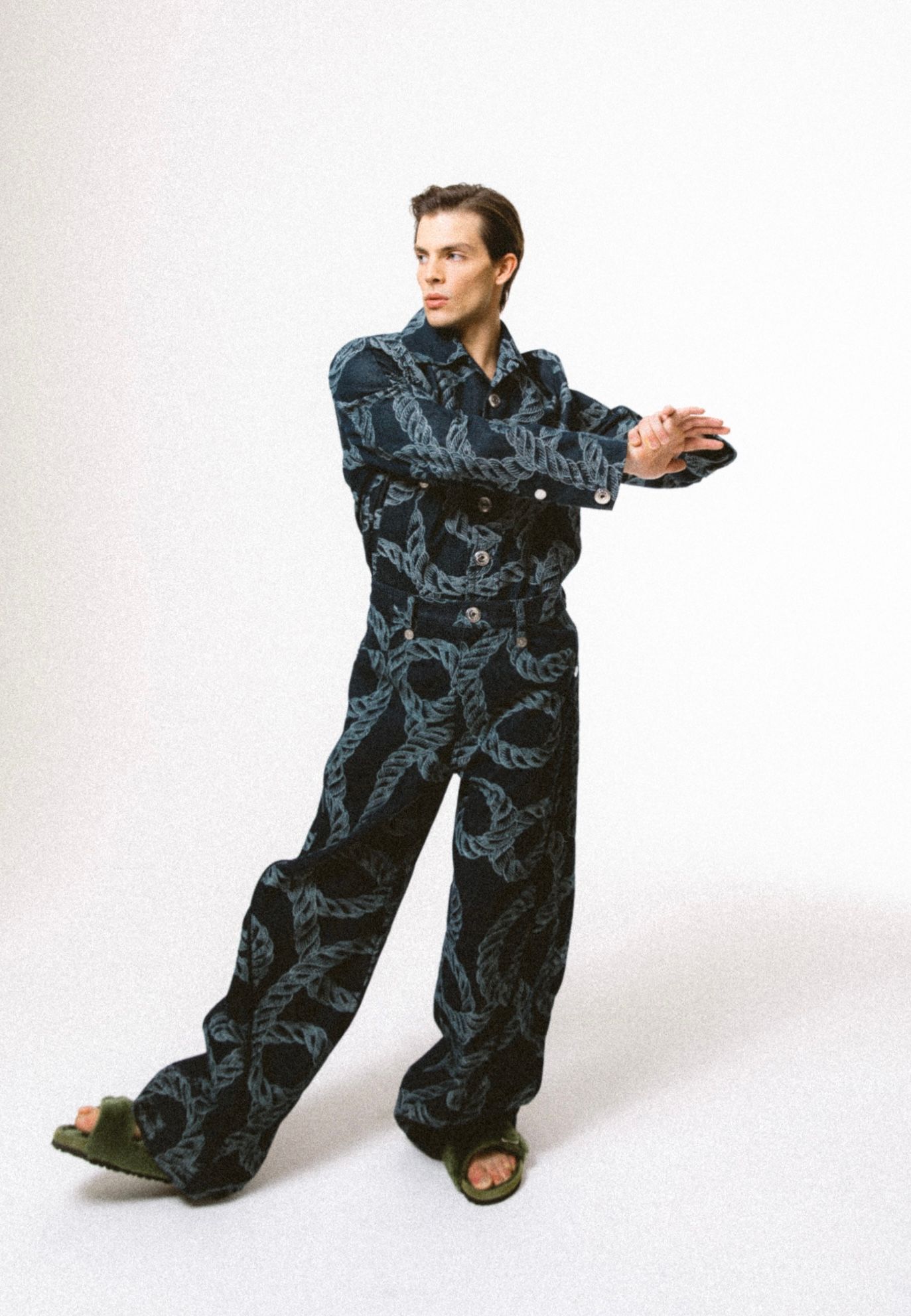 Man in patterned denim outfit, posed with arms extended, wearing sandals, on a white background.
