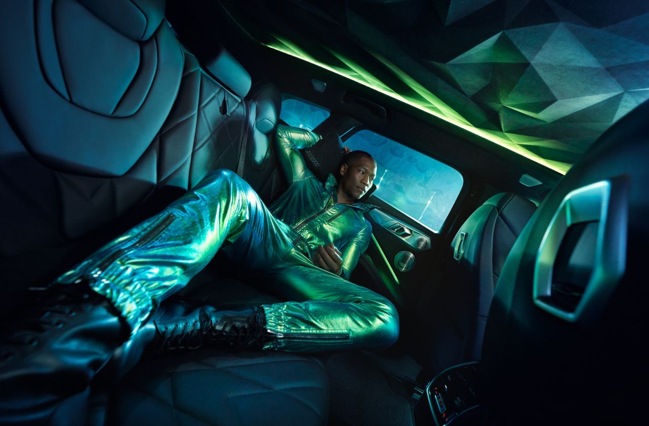 Person in a shiny, green outfit reclines in a futuristic-looking car interior with green and blue lighting.