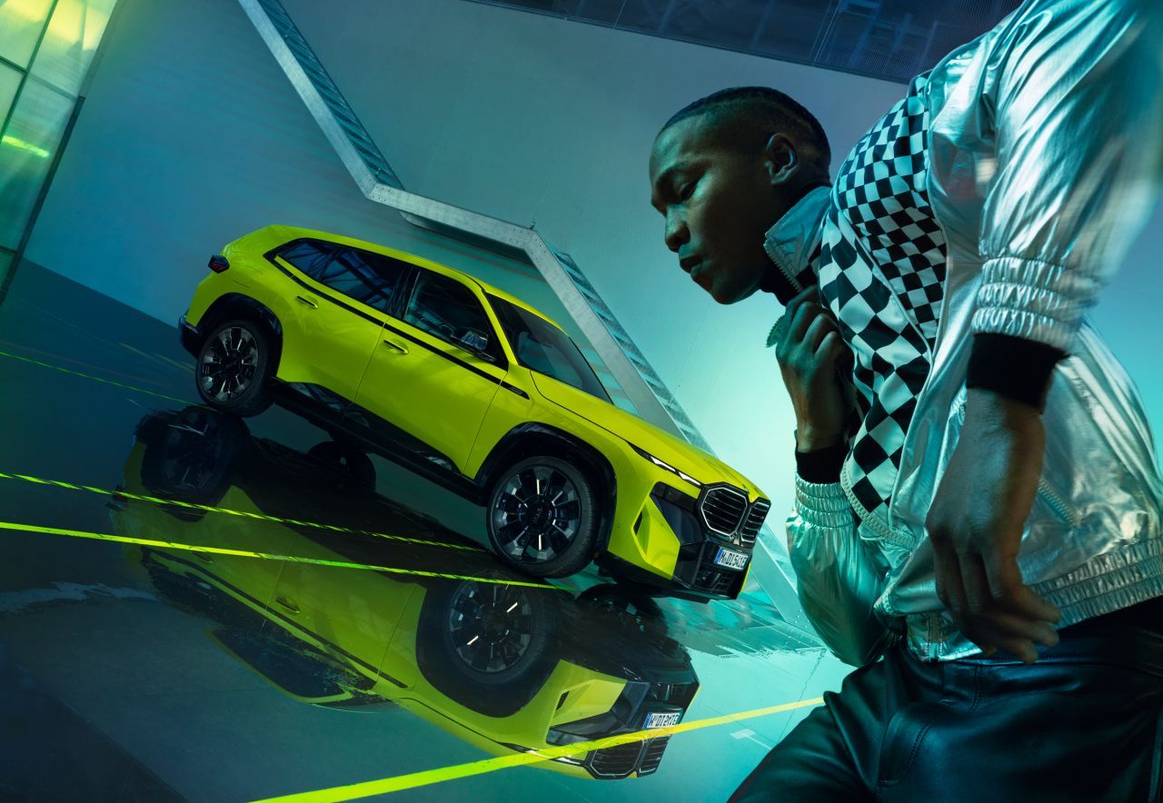 Yellow SUV alongside a person in a silver jacket and checkered shirt, with a futuristic backdrop.