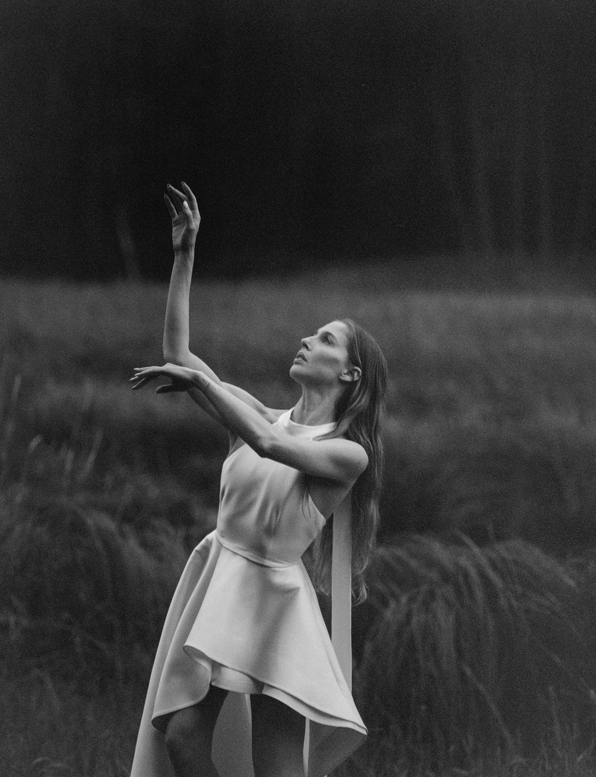 Woman in a white dress with arms raised, dancing in a field under a dark sky.