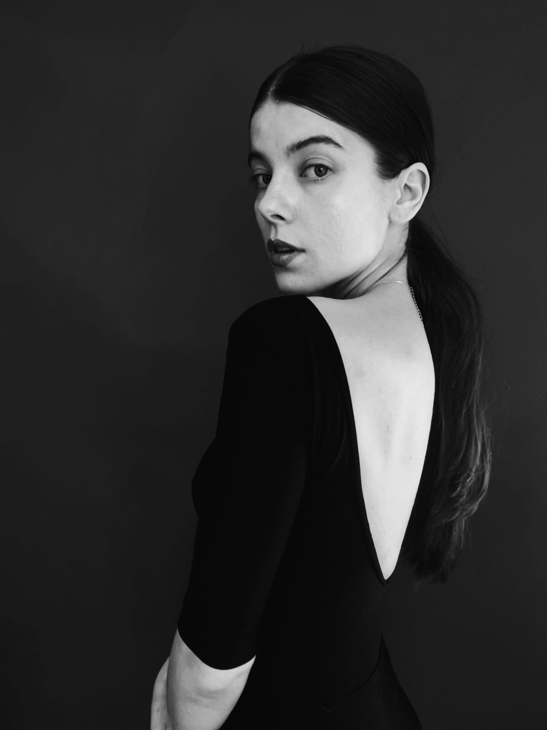 Woman in a low-back black dress, looking over her shoulder, hair in a ponytail.