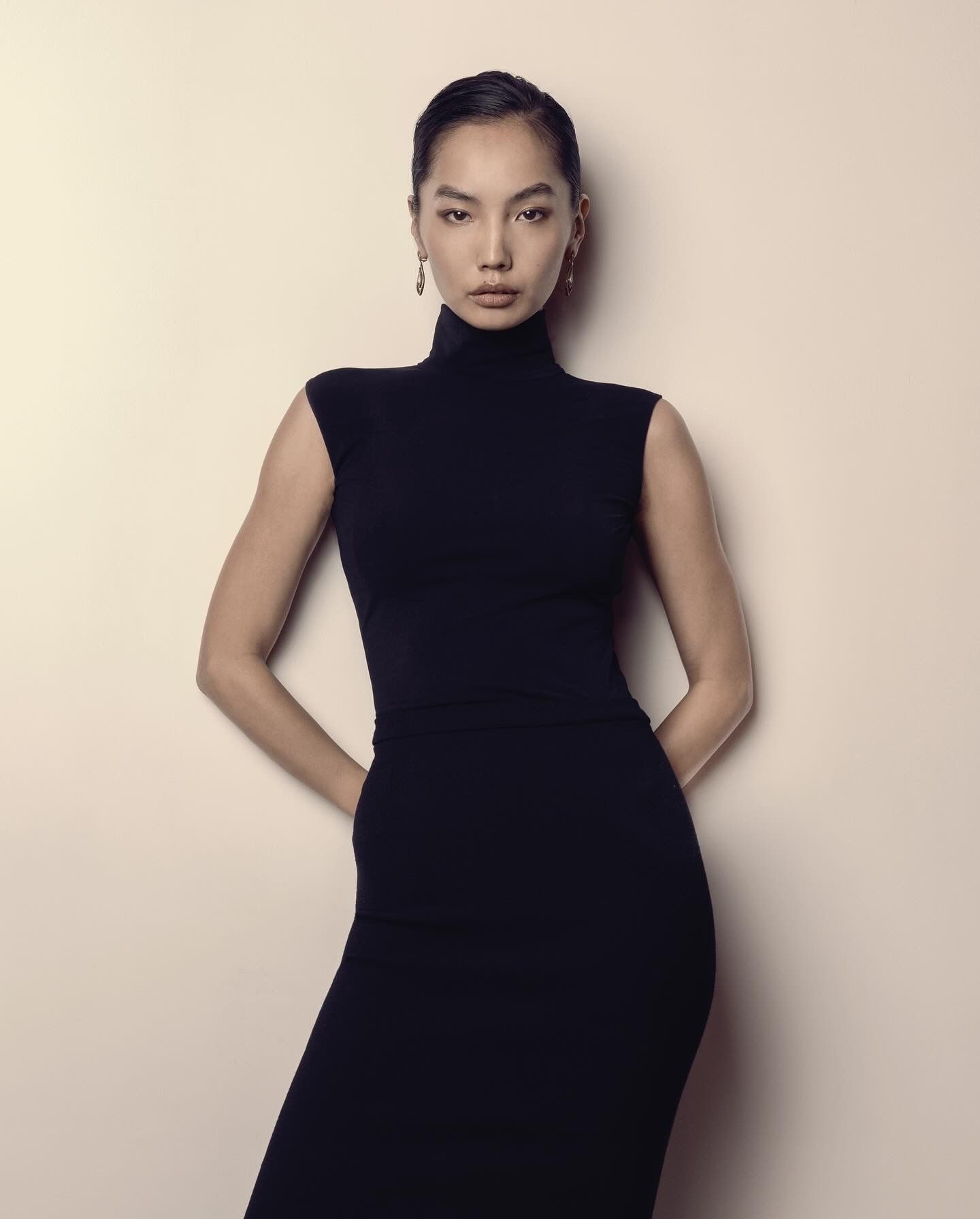 Woman in black sleeveless turtleneck dress, hands clasped behind back, leans against a light-colored wall.