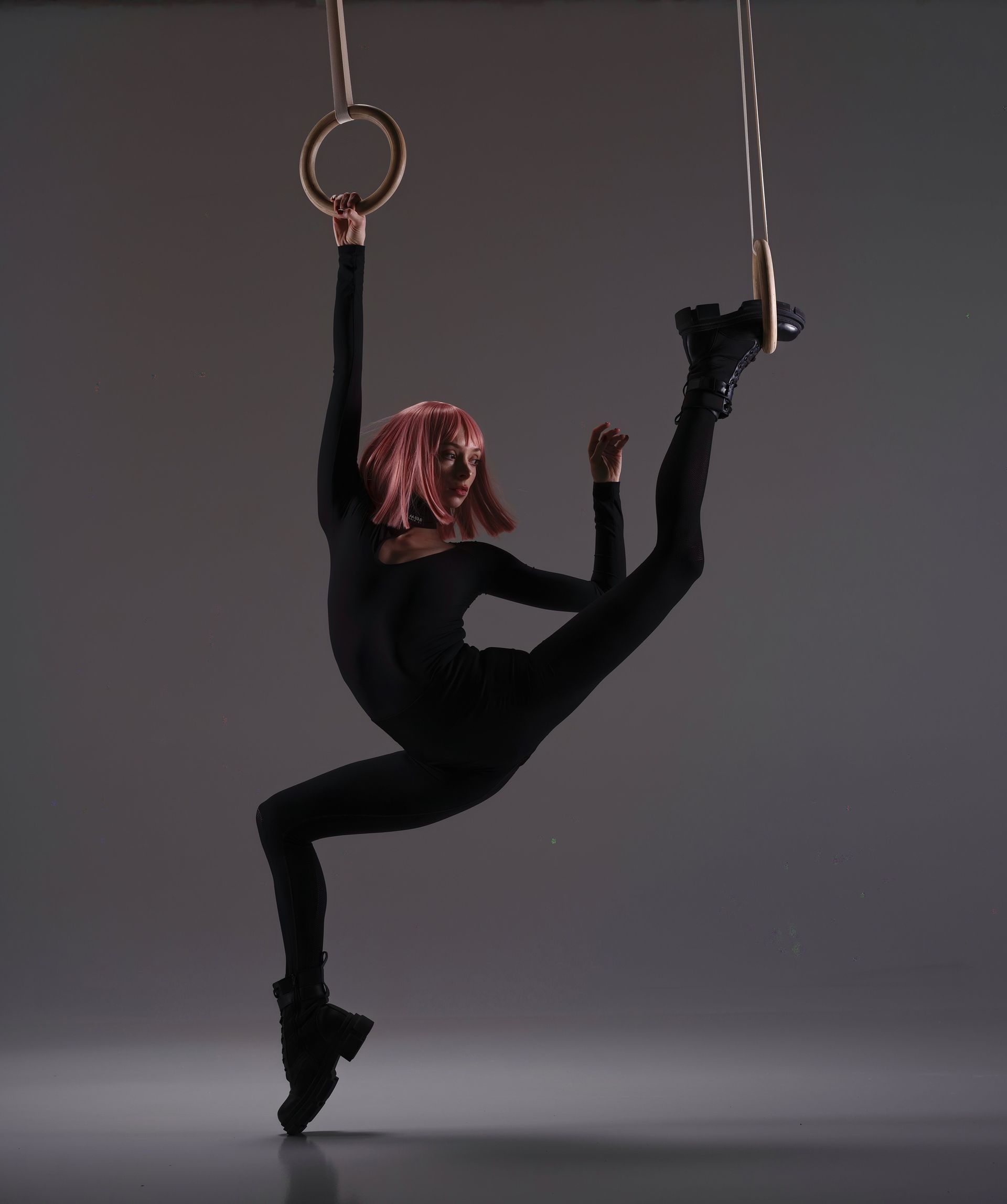 Gymnast on rings in a black leotard, boots, and pink hair, set against a gray background.