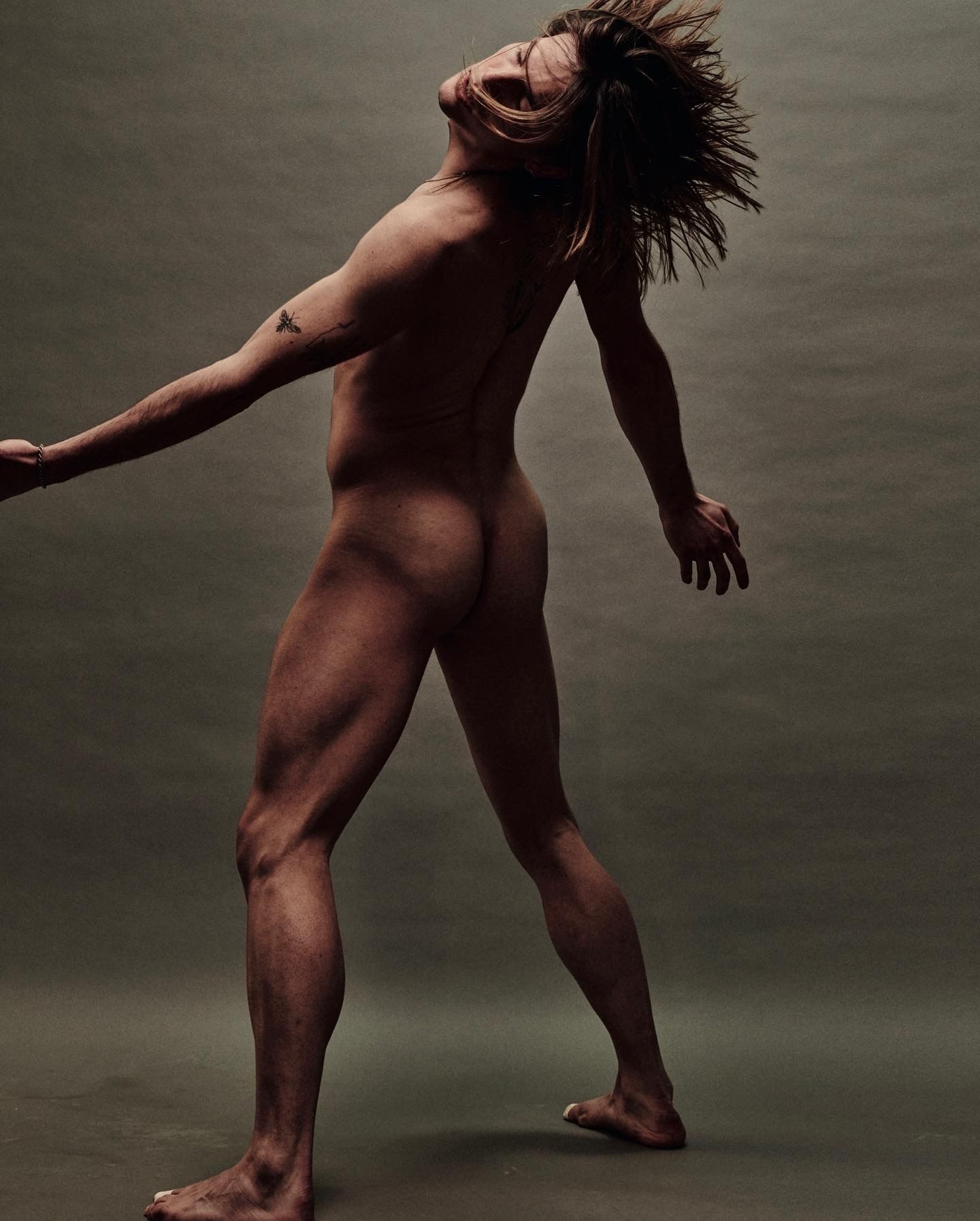 Nude person with dreadlocks arches back, arms outstretched, in front of a neutral backdrop.