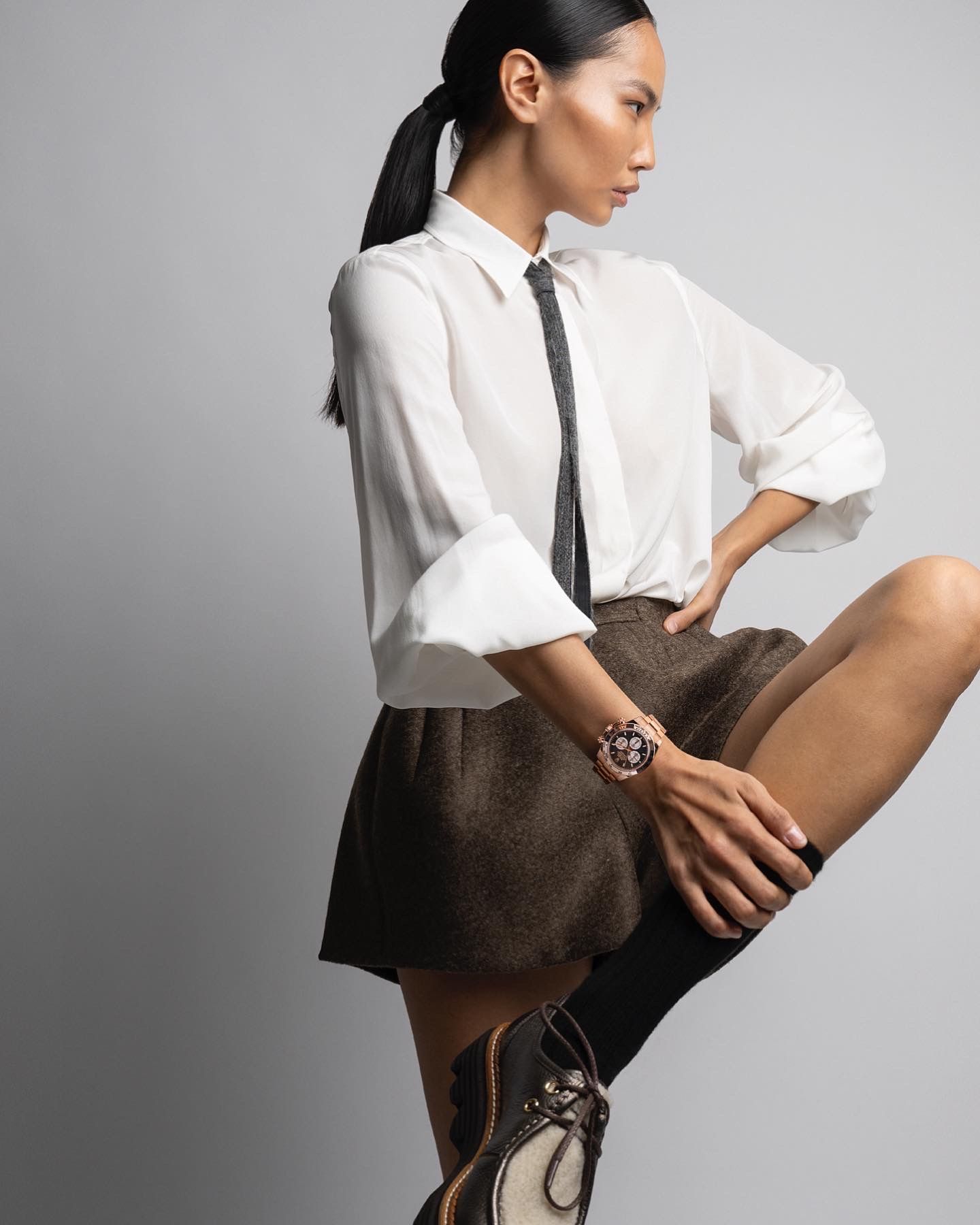 Woman in white shirt, black tie, and brown skirt, knee high boots. Looking to the side, posed with leg up.