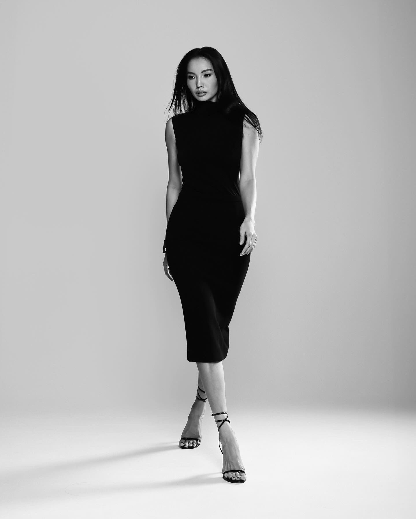 Woman in a black dress and heels, standing and posing against a neutral backdrop.