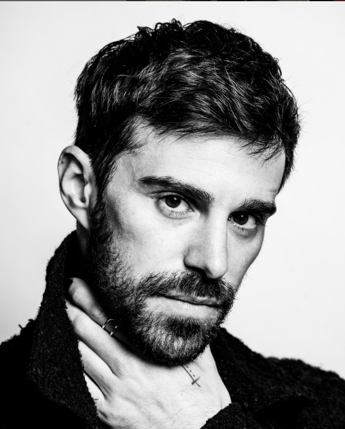 Man with dark hair and beard, looking intently at the camera, wearing a dark top, hands near his neck.