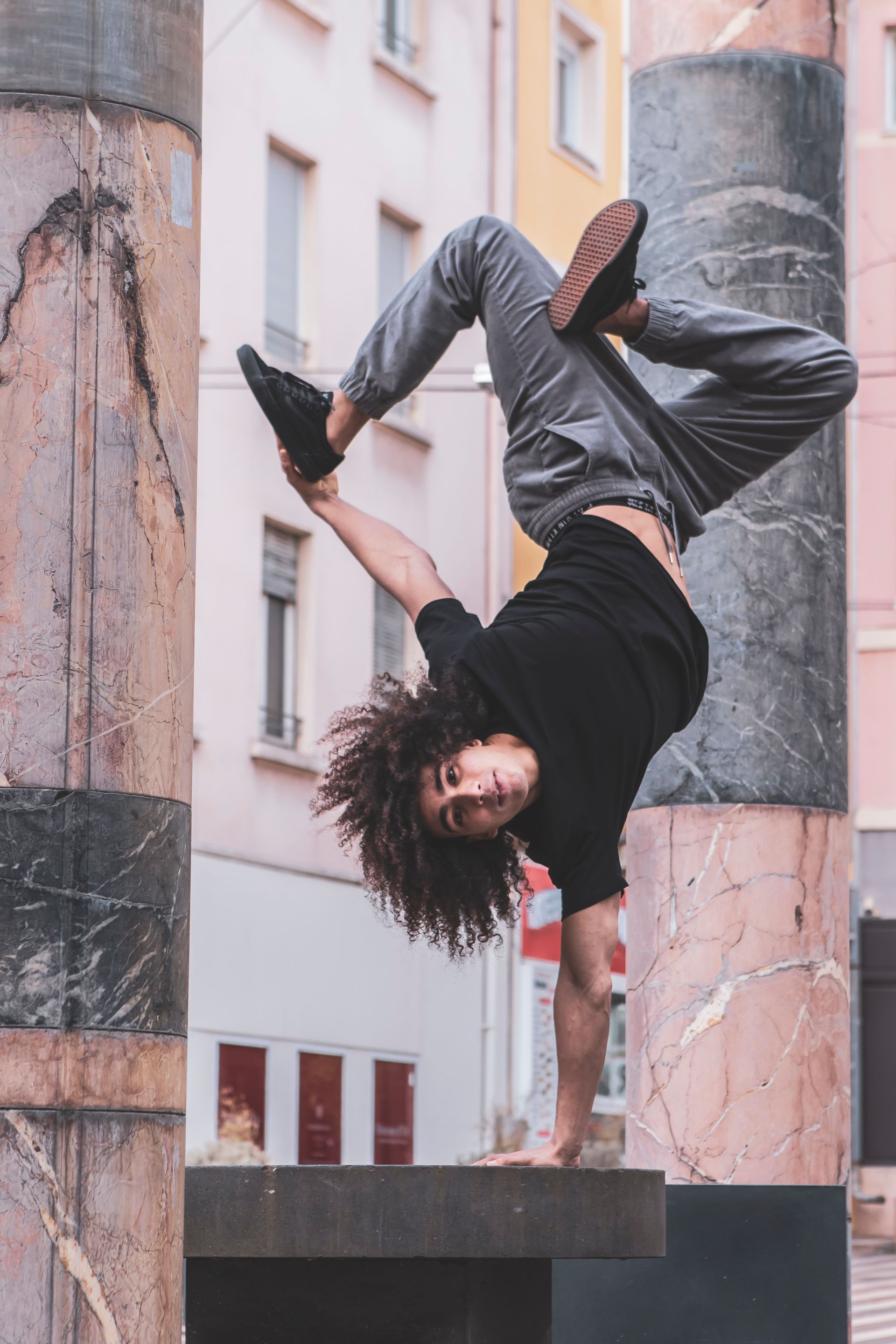A man balances upside down on one hand, legs bent, in front of pillars and buildings.