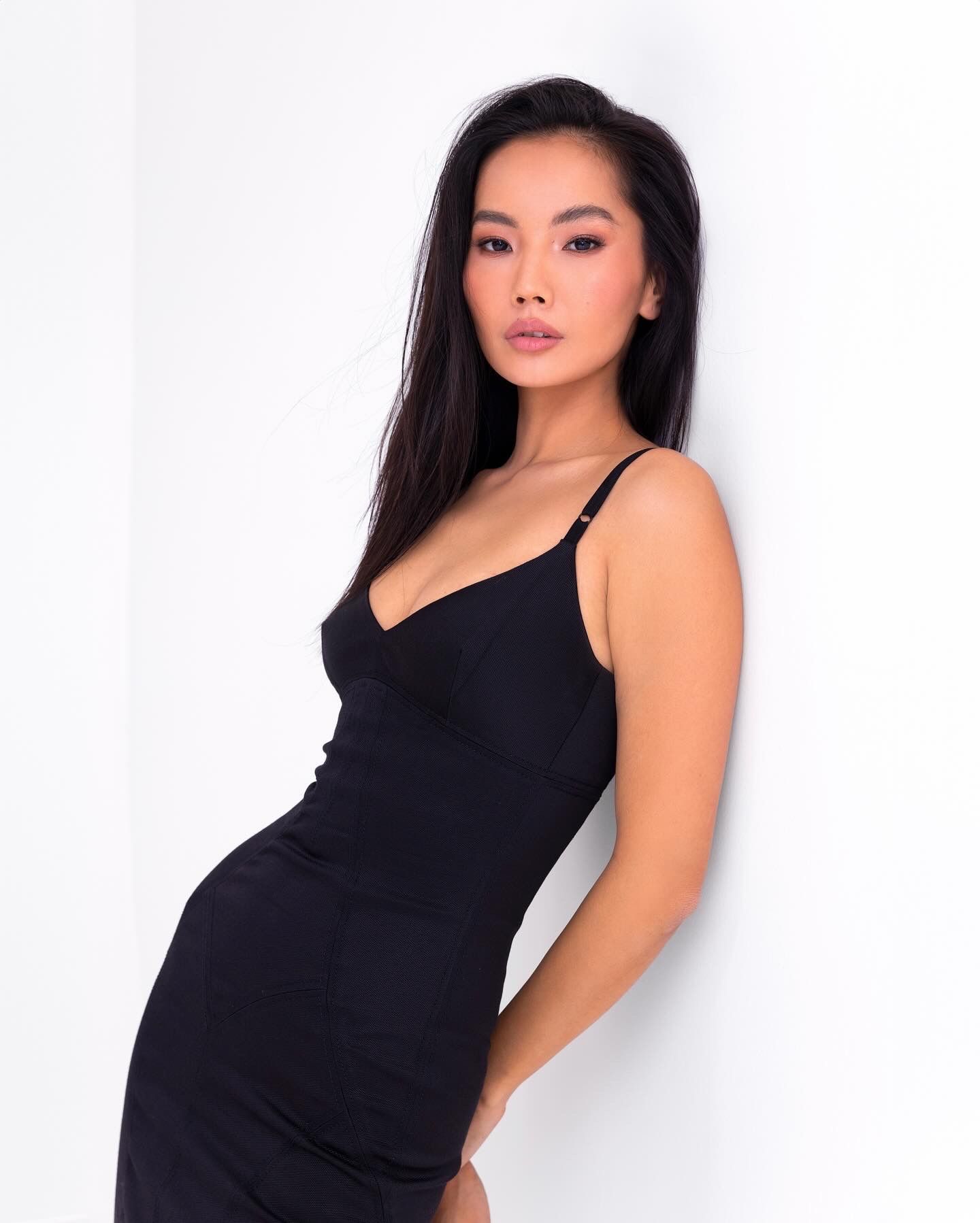 Woman in a black dress leans against a white wall, looking at the camera.