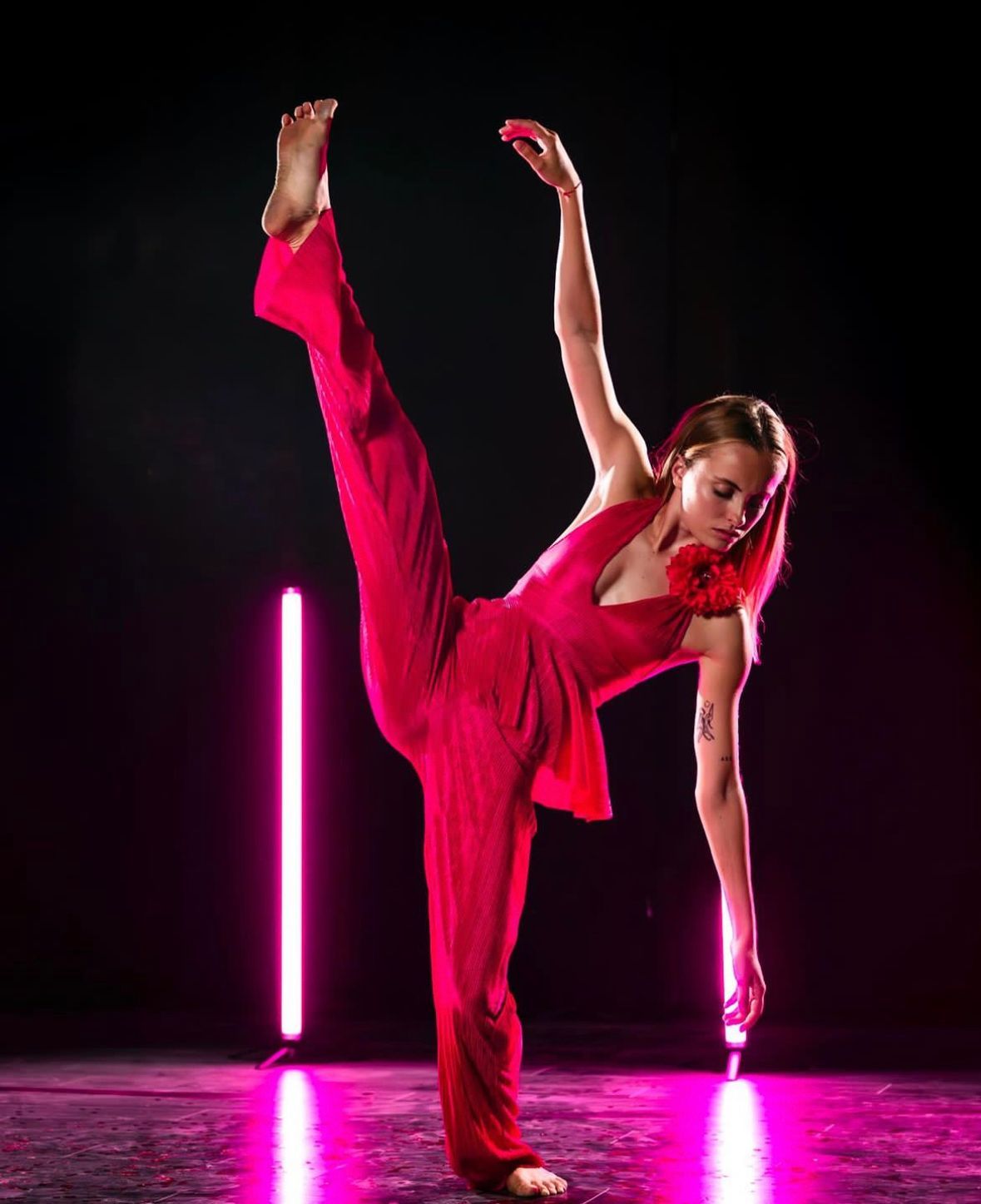 Dancer in a red jumpsuit, leg extended high, arm raised, performing in front of pink neon lights.