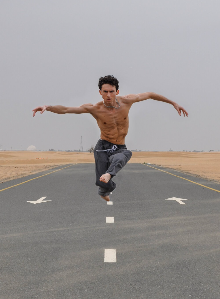 Man mid-air on runway, arms outstretched, shirtless, wearing dark pants. Desert background, overcast sky.