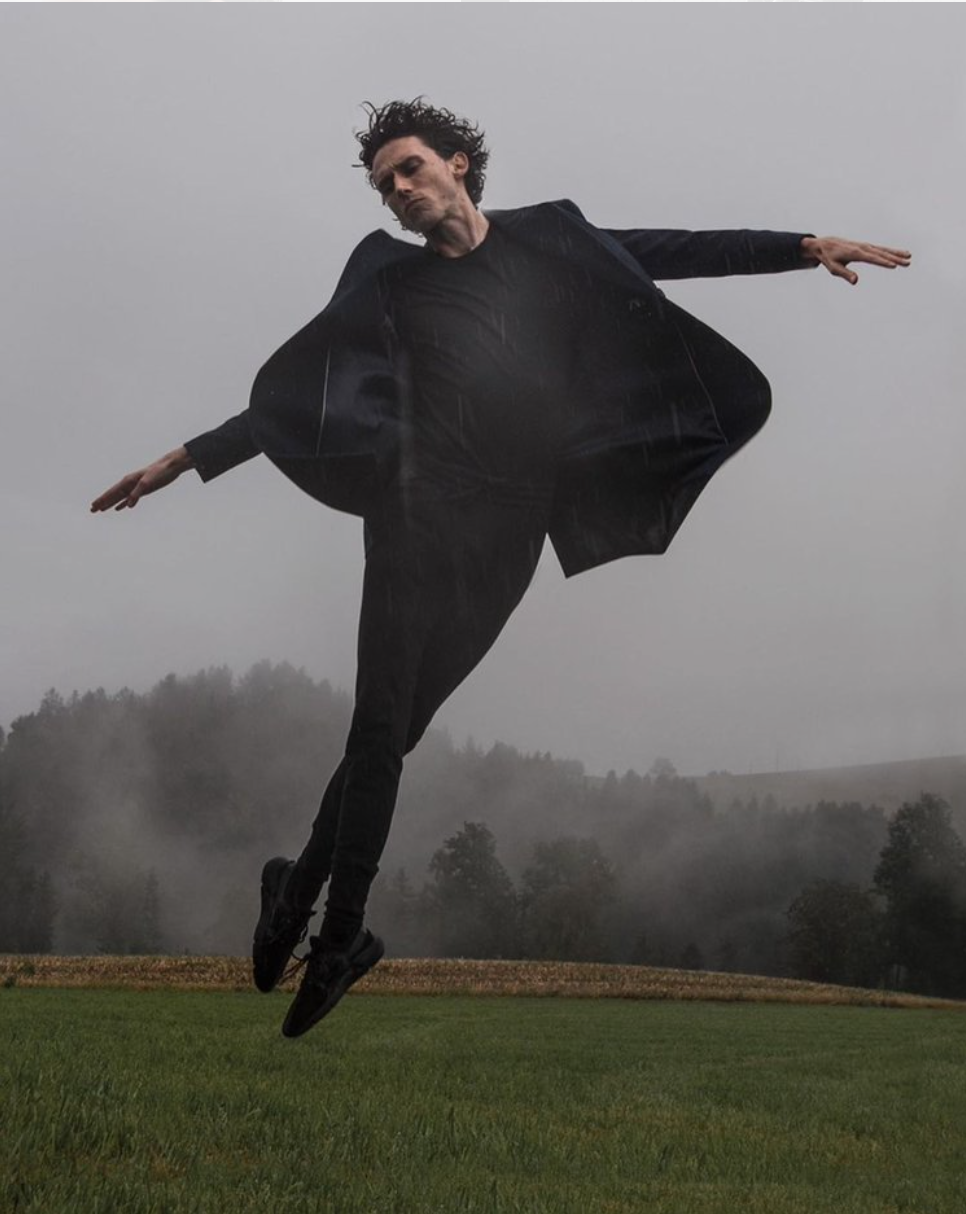 Man in black suit levitating above green grass, arms outstretched, misty forest background.