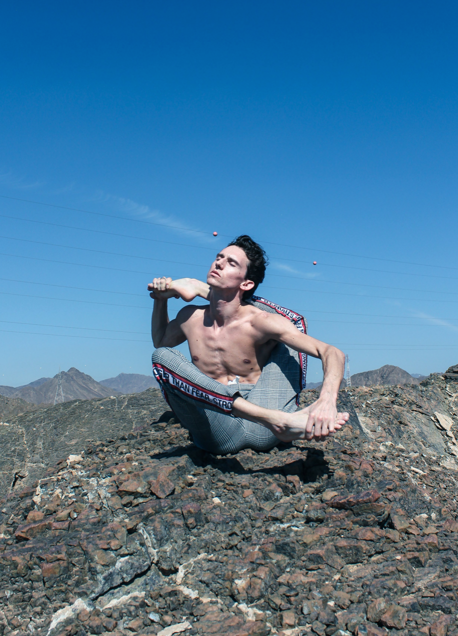 Man contorted in a yoga pose on a rocky hilltop, shirtless, holding his foot. Clear blue sky.