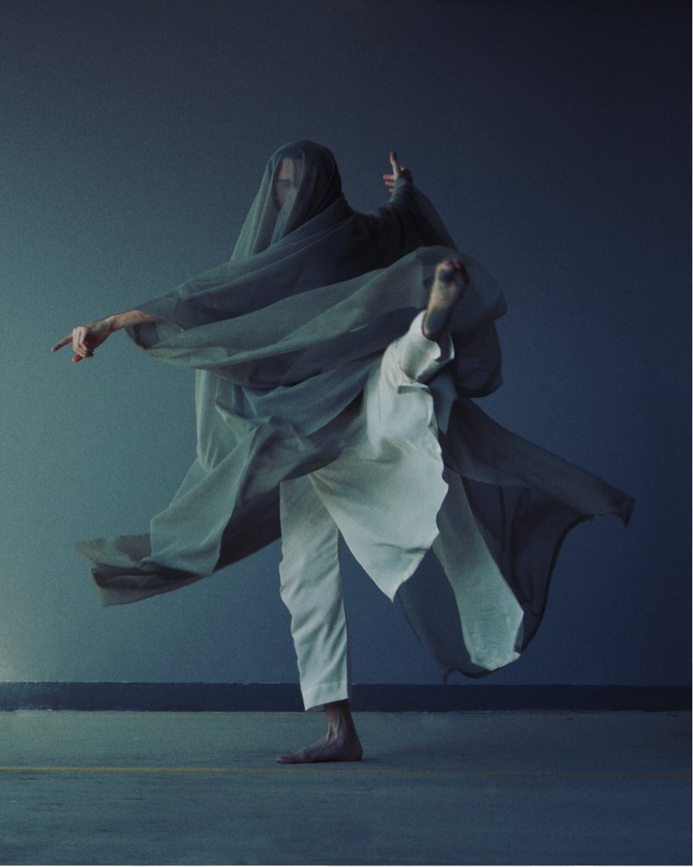 Person in flowing fabric, balanced pose, arms outstretched. Against a blue-grey wall.